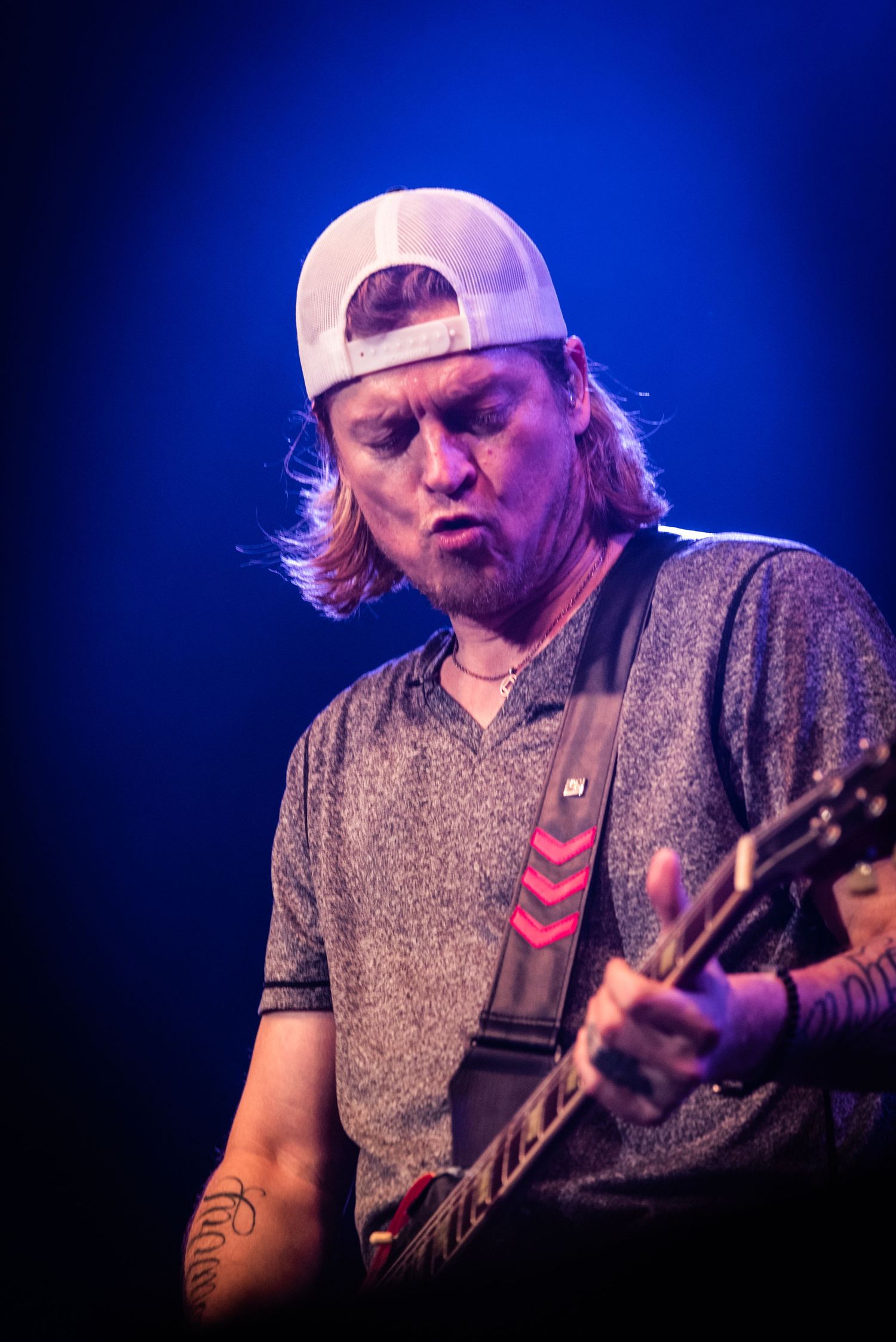Guitarist wearing backwards cap and gray shirt plays intensely under blue stage lighting.