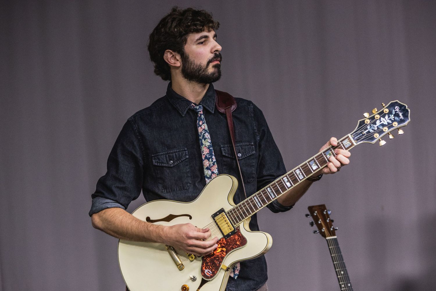 Guitarist playing cream-colored semi-hollow body guitar on stage with purple backdrop.