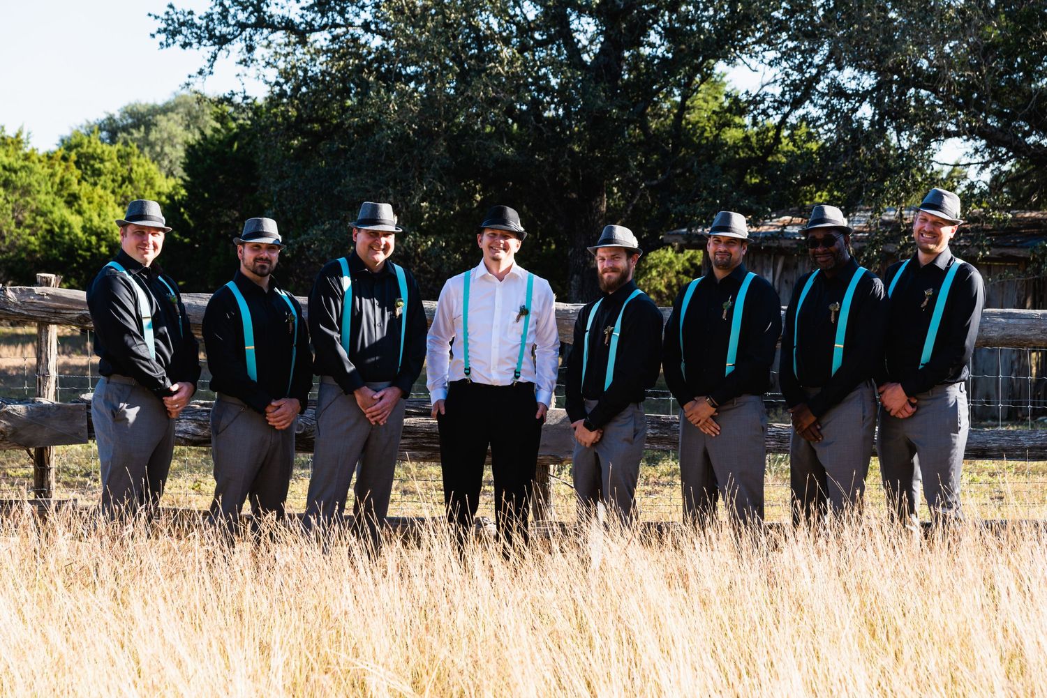 Wedding party in black suits with turquoise suspenders stands together in golden grass field.