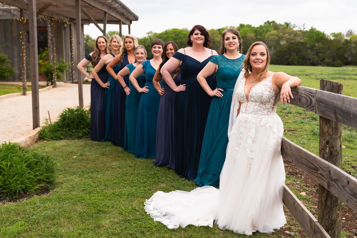 Wedding party in teal and navy dresses posed along rustic fence with bride in white gown.