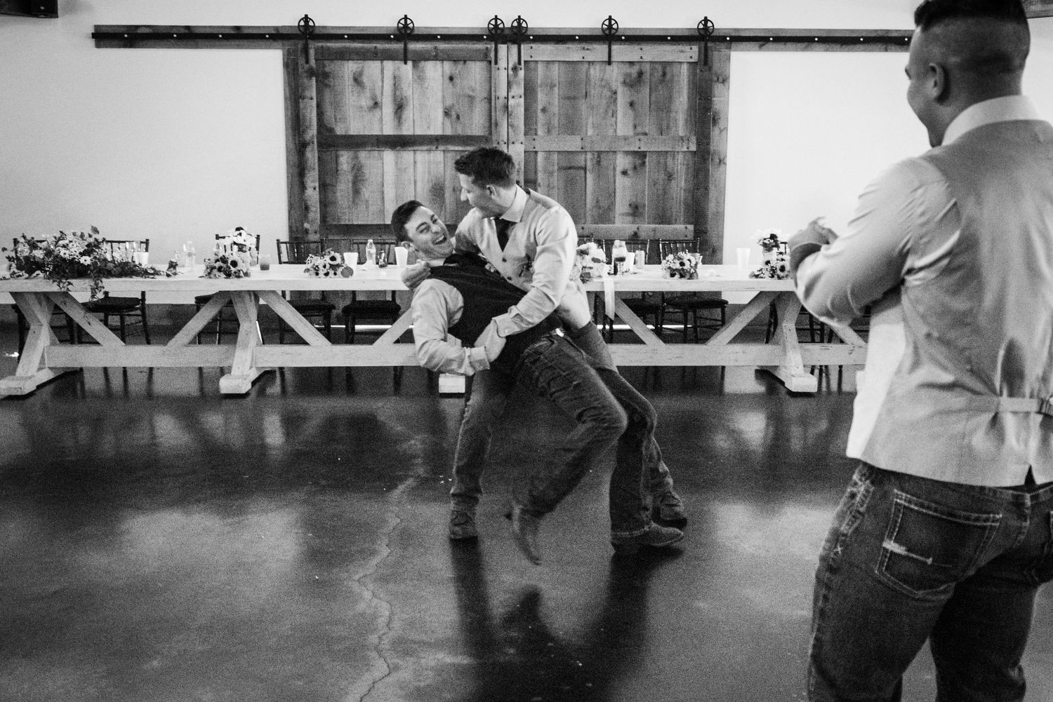 A black and white photo captures energetic dancing at a rustic reception venue.