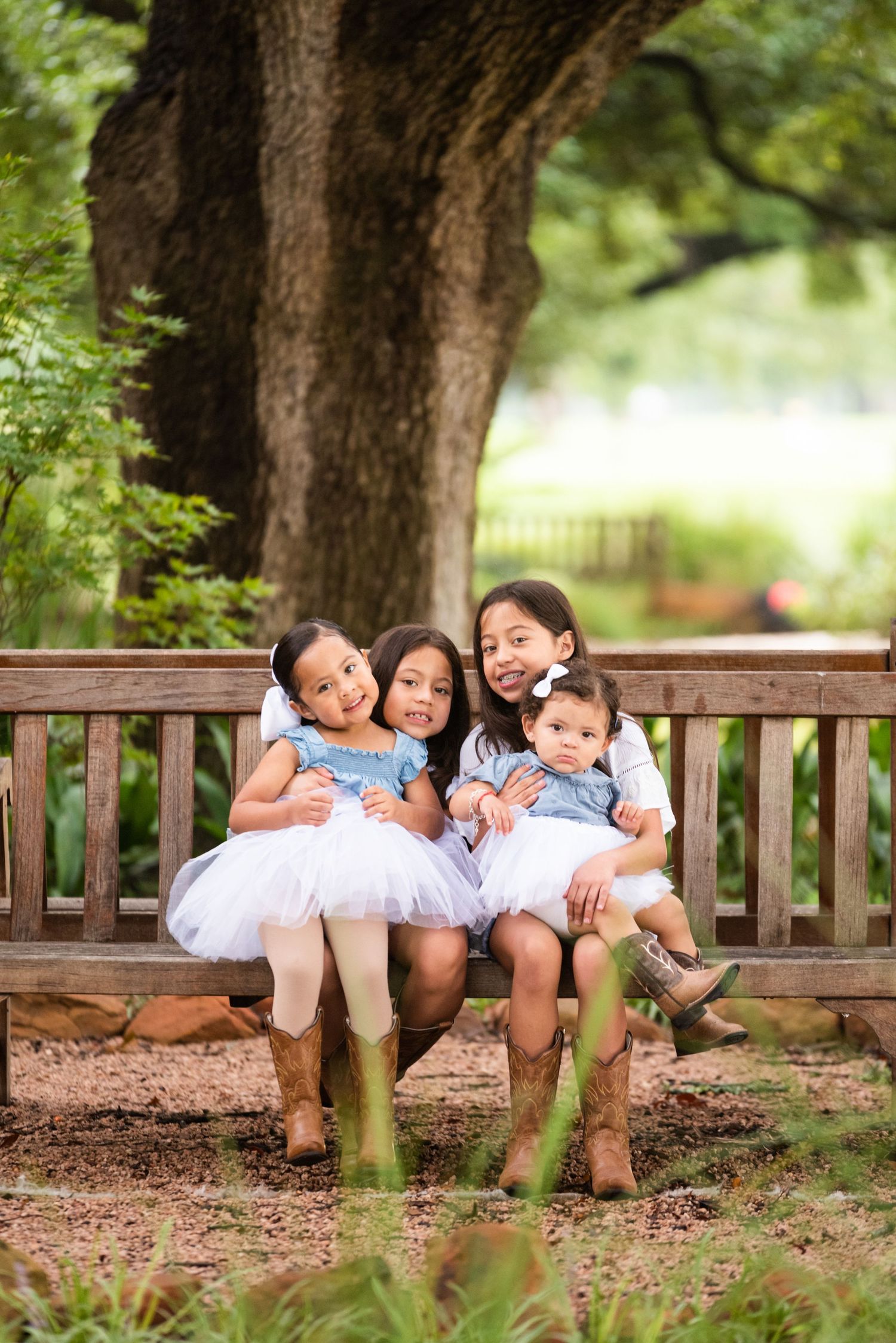 Children in white dresses and boots sit together on a wooden bench under a tree.