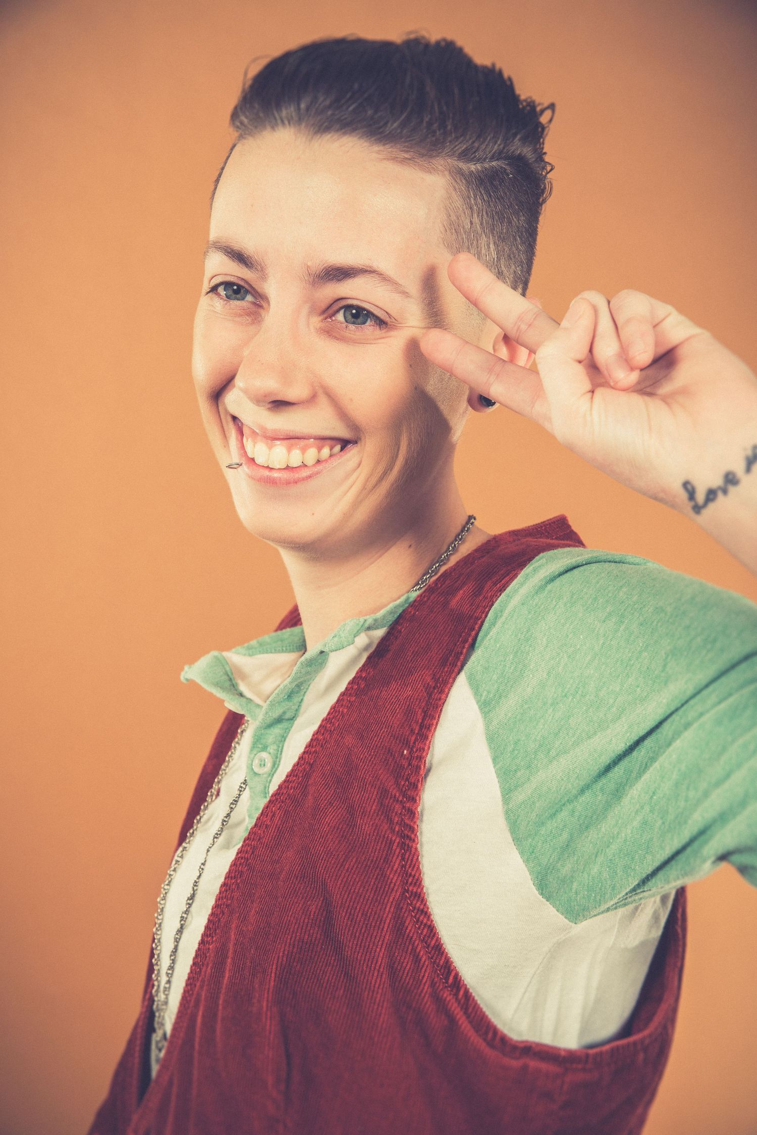 Portrait of person in vintage vest making peace sign with bright smile against orange background.