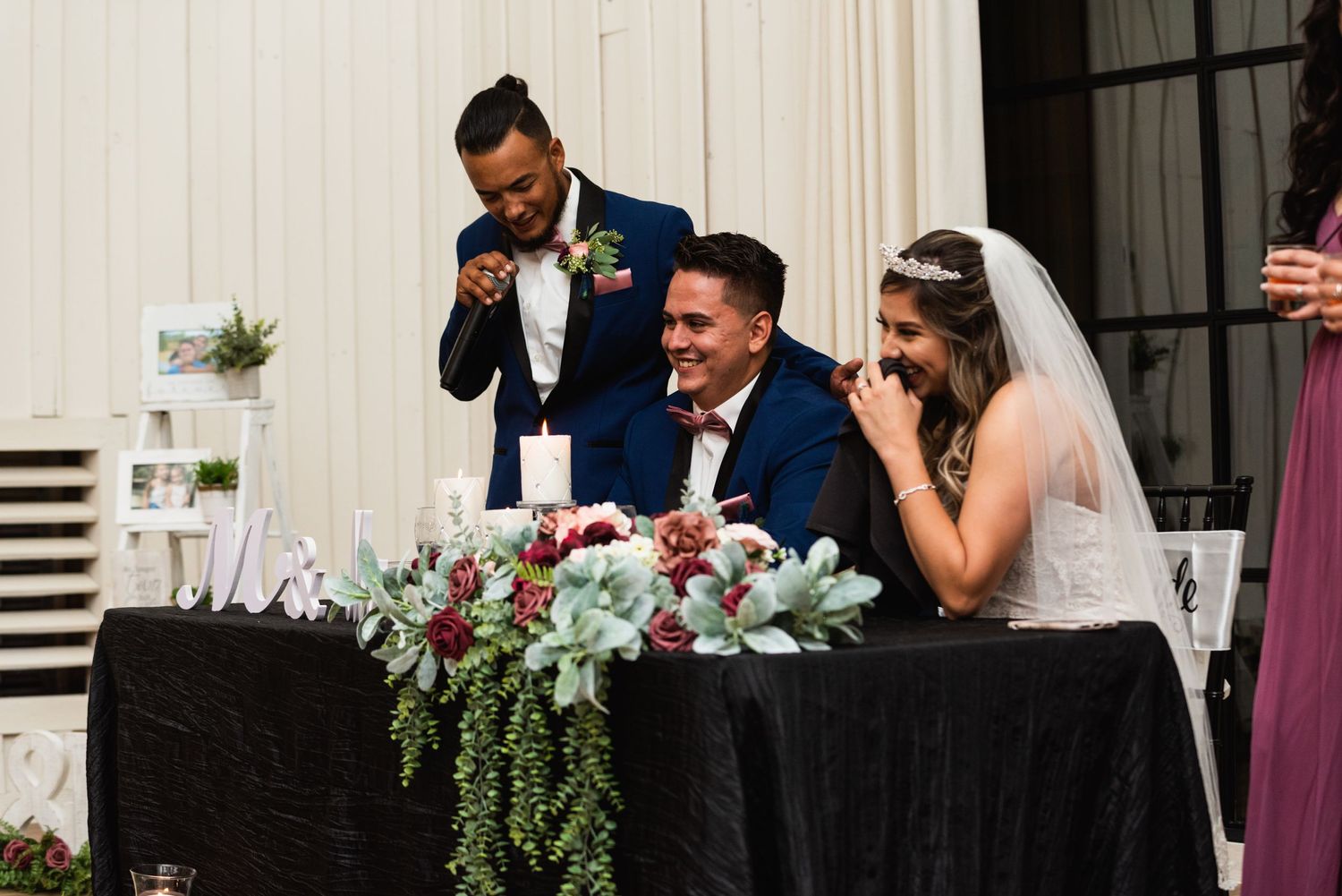 Wedding party seated at head table with floral arrangements during reception speeches.