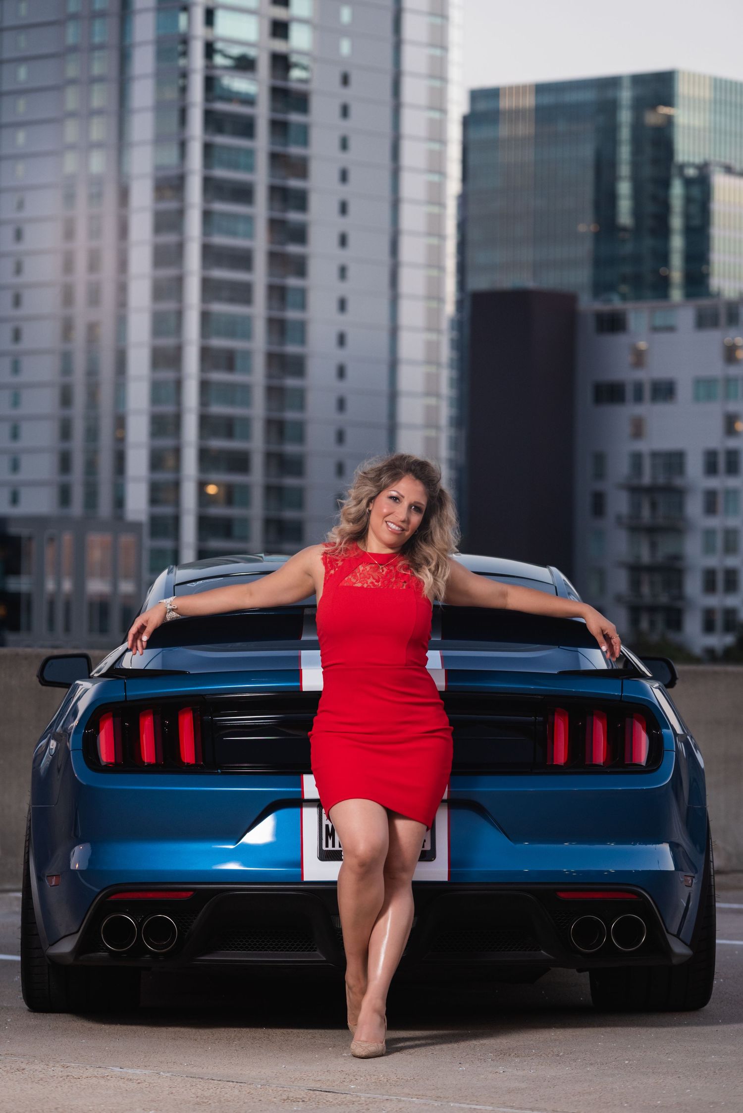 Stylish red dress contrasts with blue Mustang against city skyline backdrop.