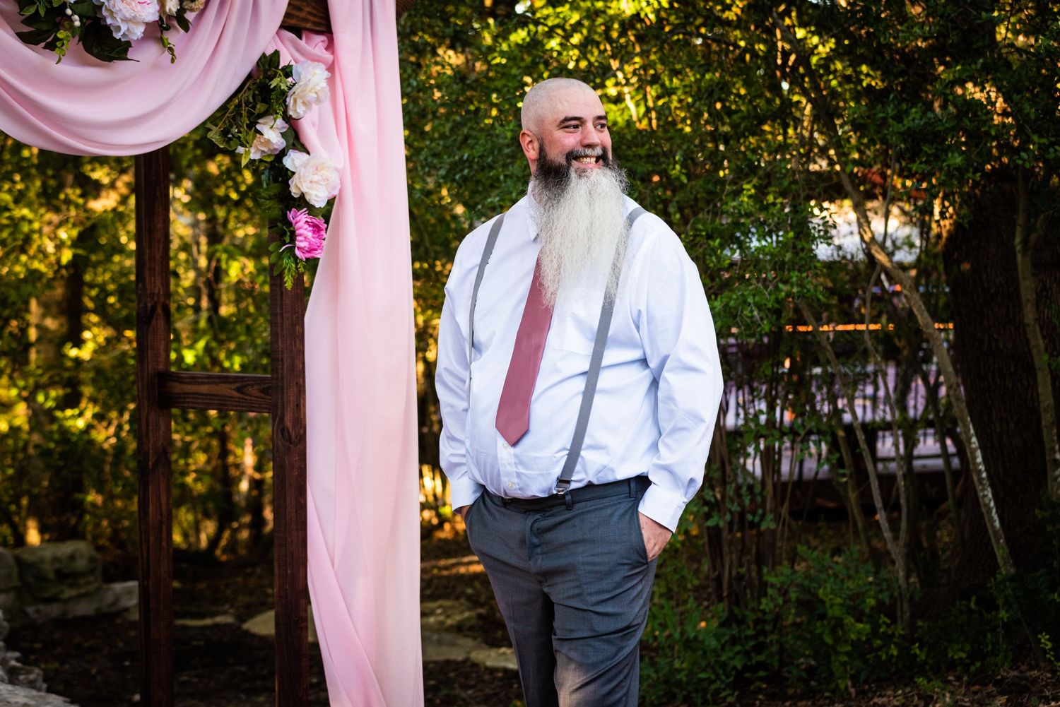 A person in formal attire with a pink tie stands near pink wedding decorations outdoors.