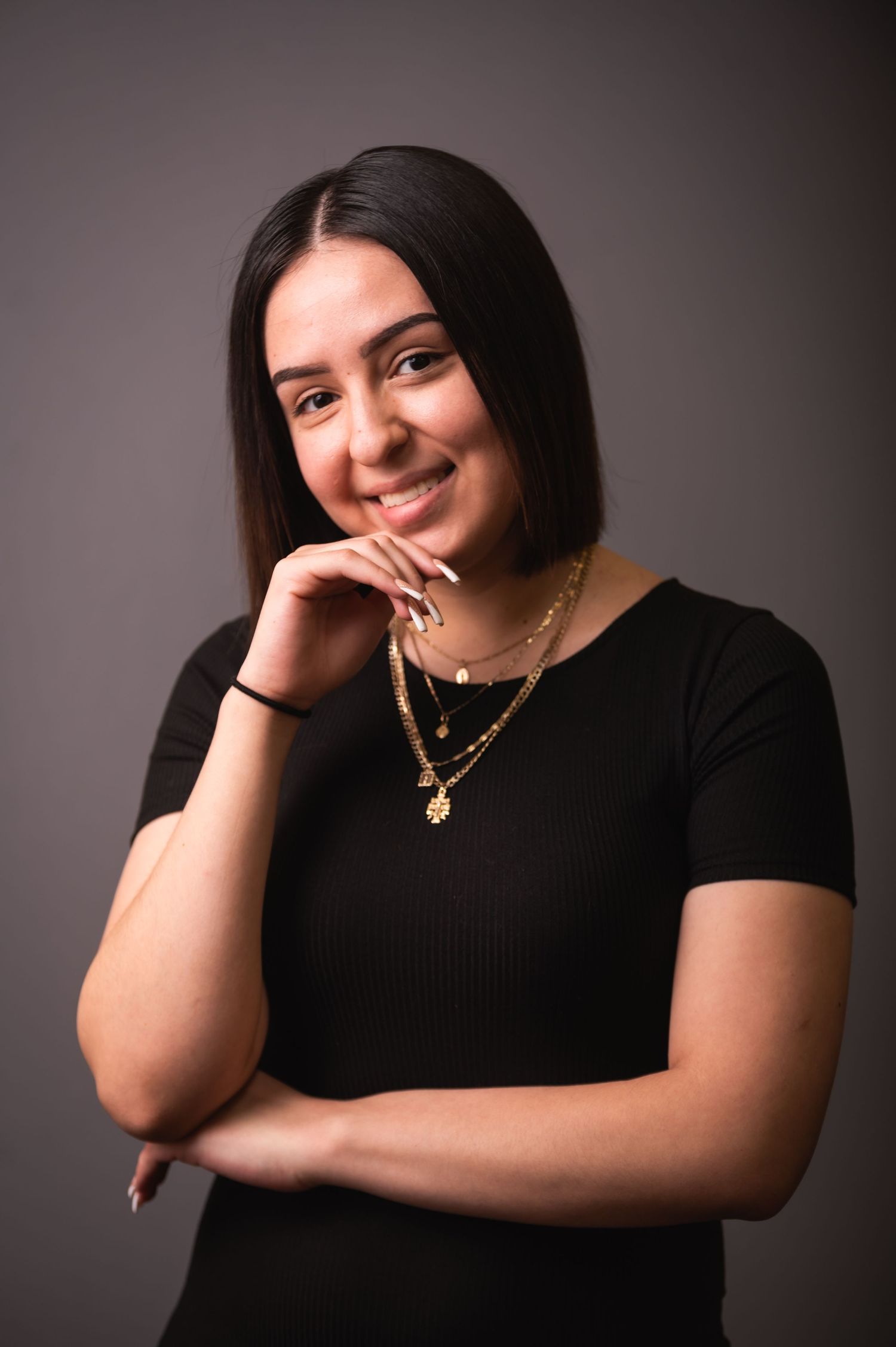 Portrait of person in black top with layered necklaces against grey background.