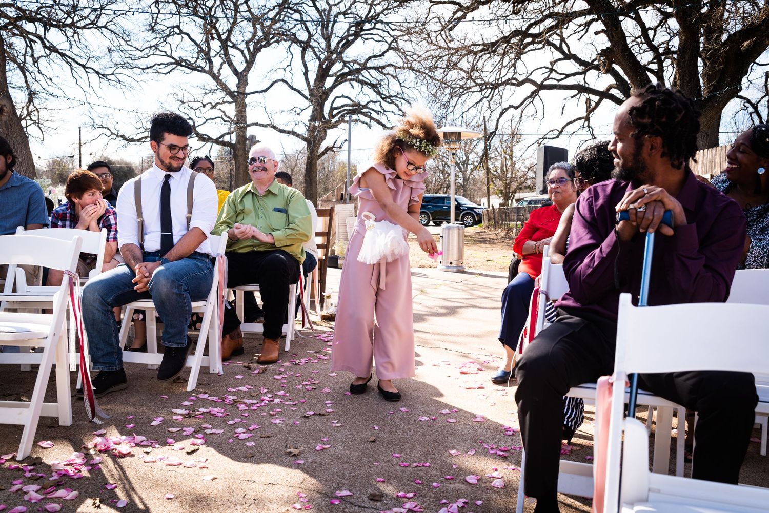 Rose petals scatter the ground during an outdoor wedding ceremony with white folding chairs.