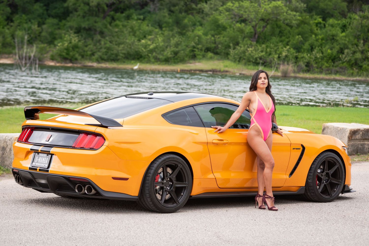 Bright orange Ford Mustang GT parked by a lake with trees in the background.