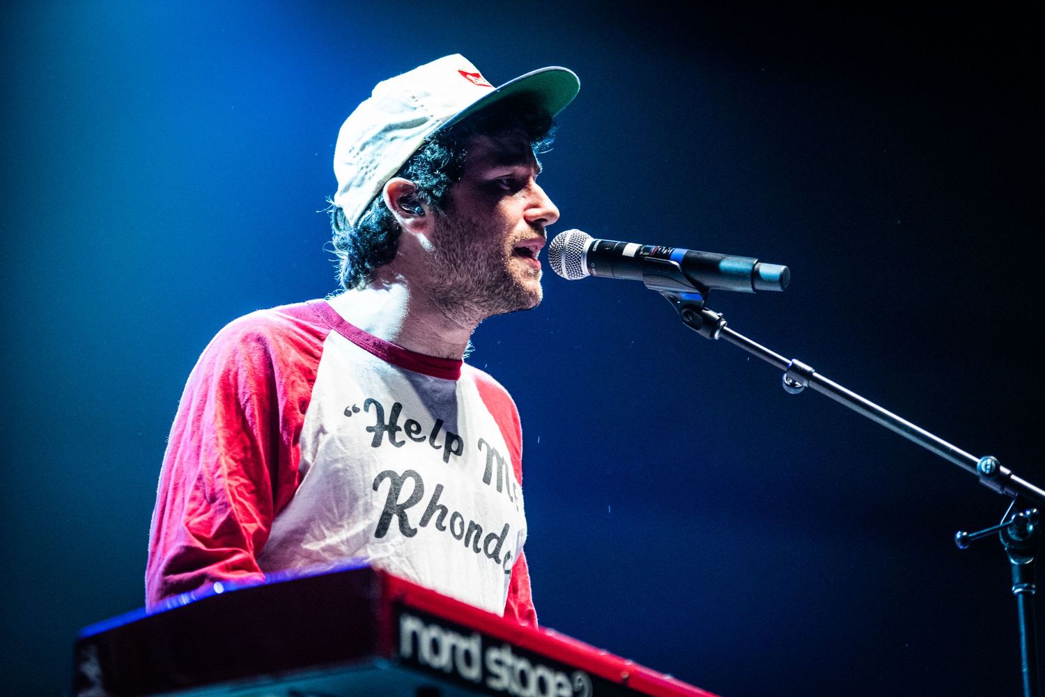 Keyboard player in baseball cap and raglan shirt performs with dramatic blue stage lighting.