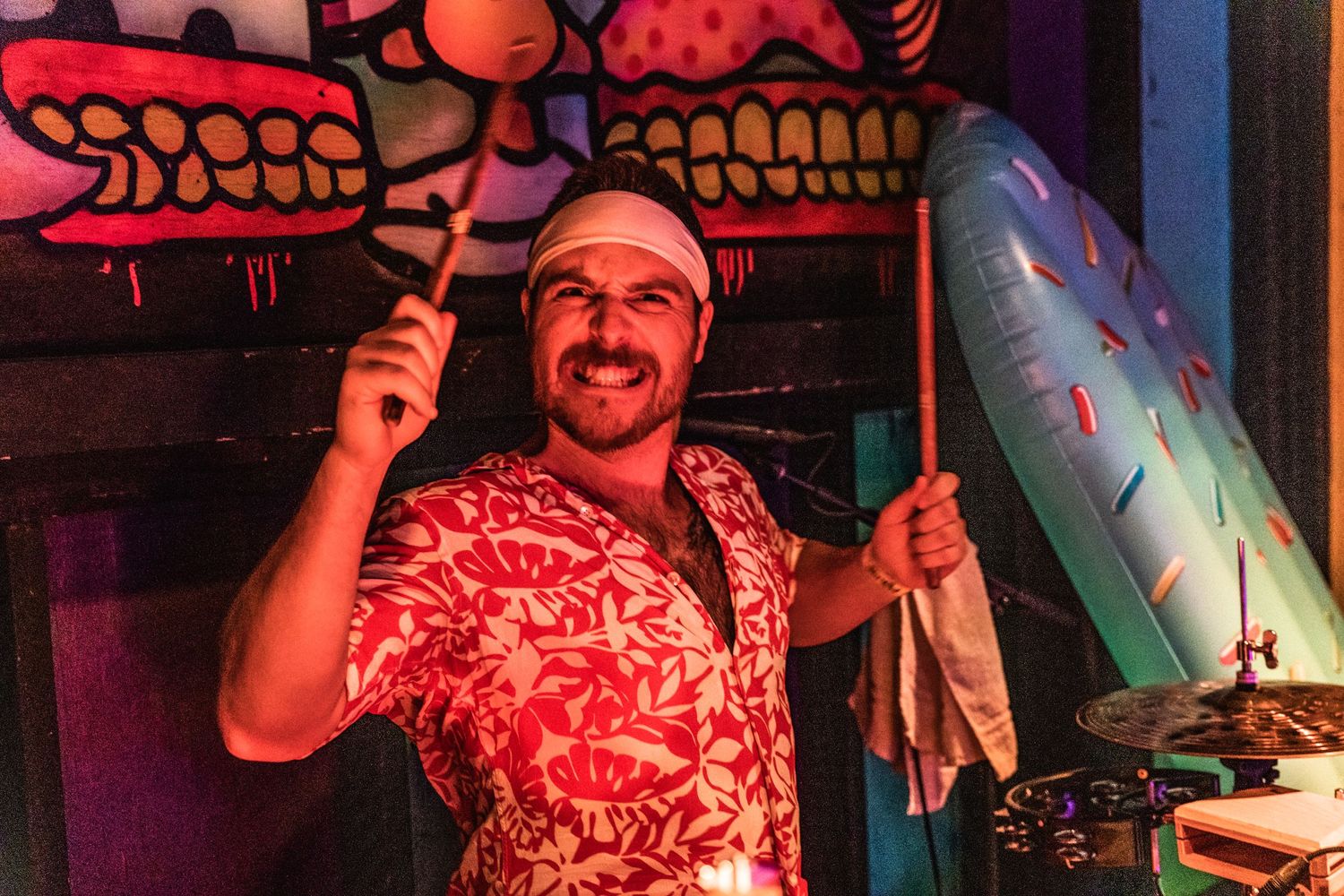 A person in a festive red Hawaiian shirt smiles while holding party decorations in a dimly lit setting.