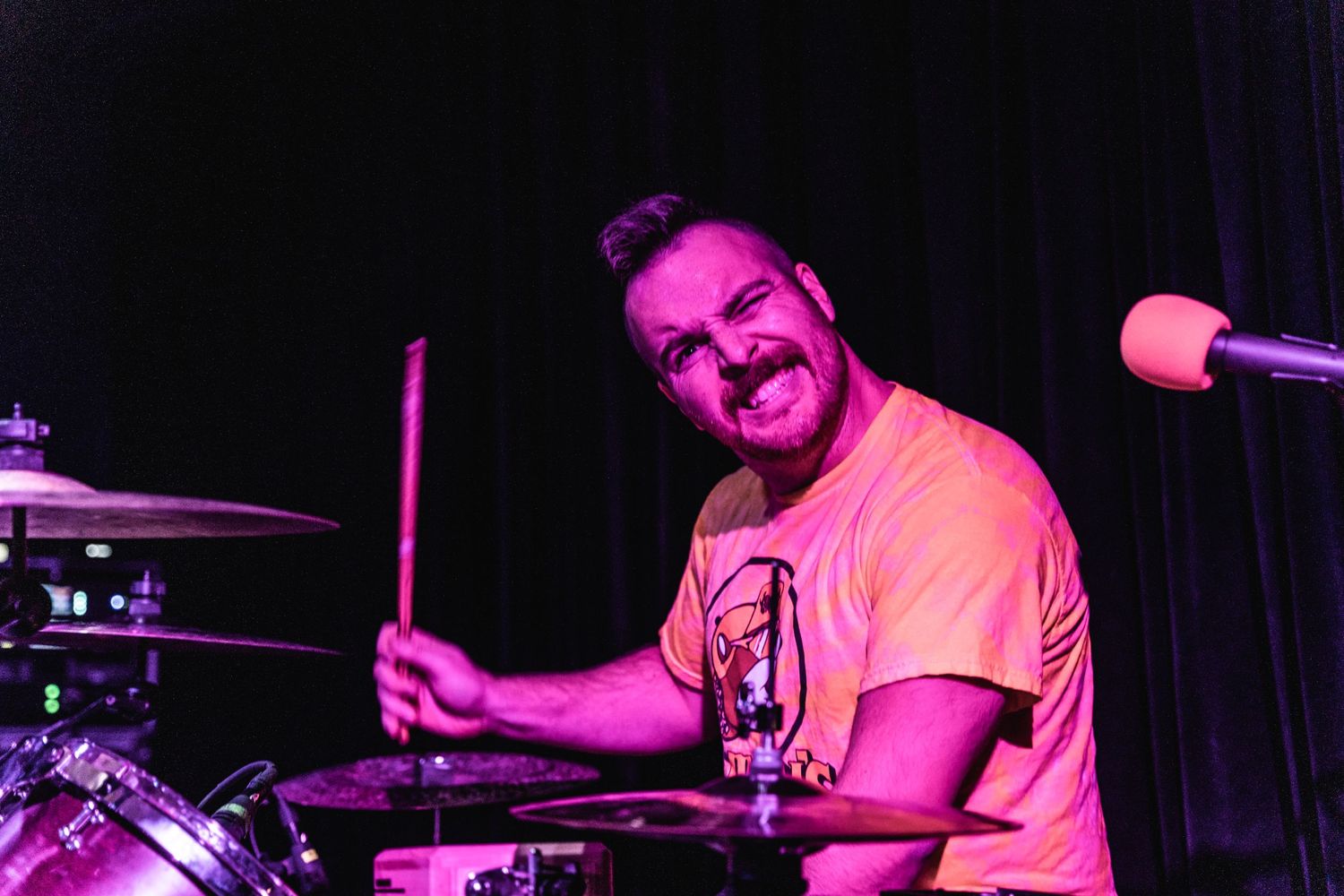 Drummer performing on stage illuminated by pink and purple lighting.
