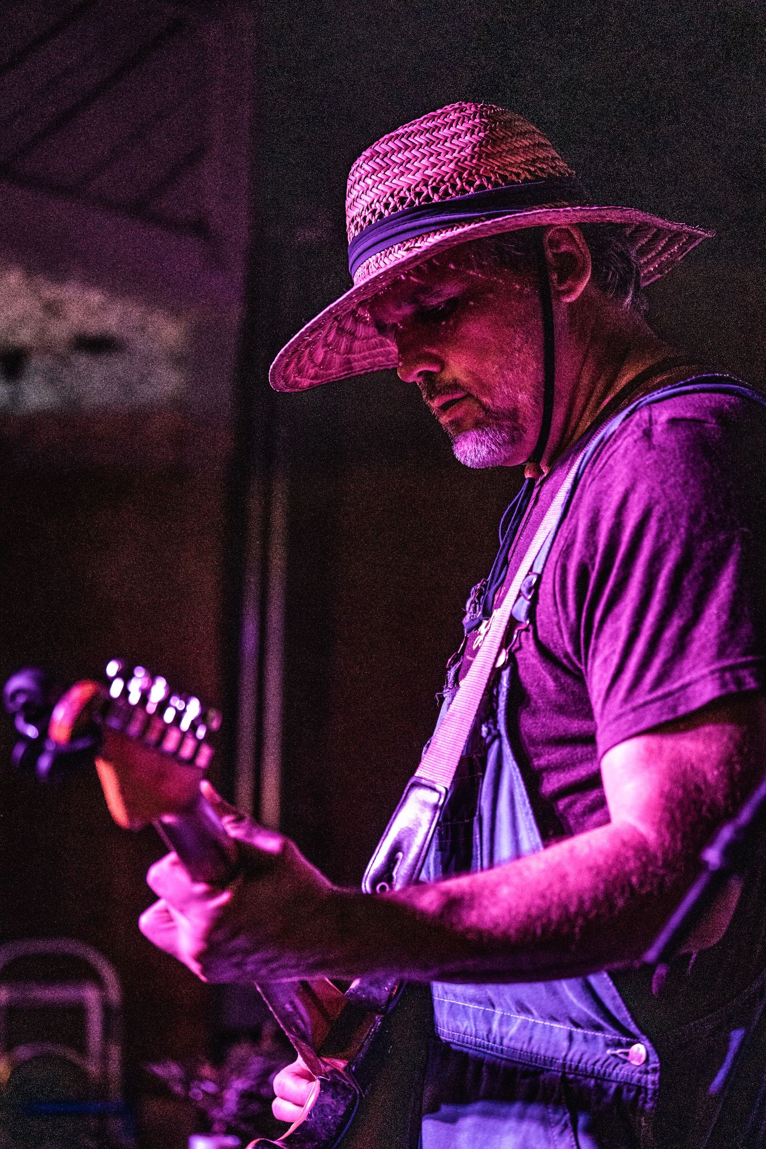 Guitarist wearing straw hat and overalls performs under dramatic pink stage lighting.