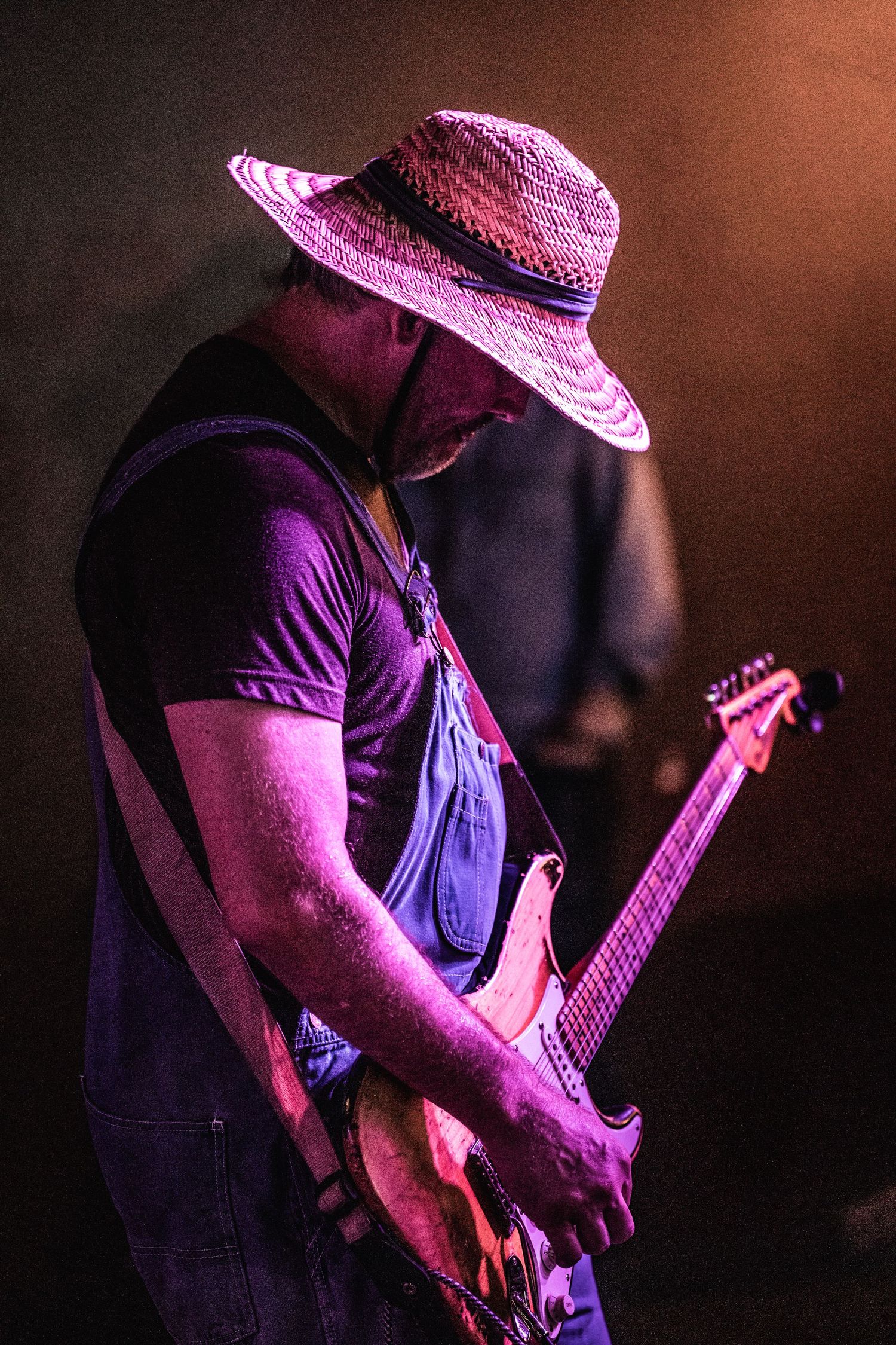 Guitar player wearing a straw hat and overalls performing under magenta stage lights.
