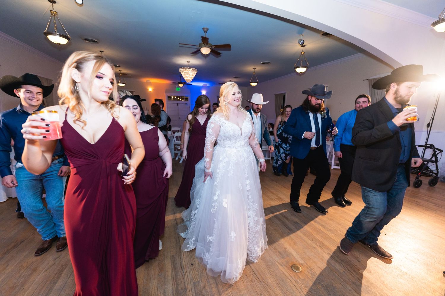 Wedding guests dance and celebrate on a wooden dance floor with blue lighting and decorative ceiling details.