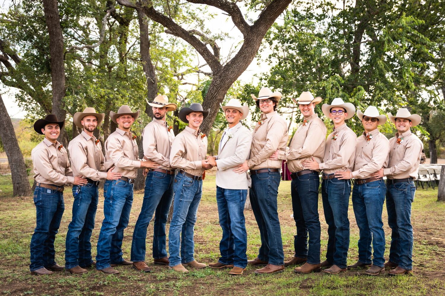 A group of groomsmen in matching light colored shirts and blue jeans pose in line under a large tree outdoors.