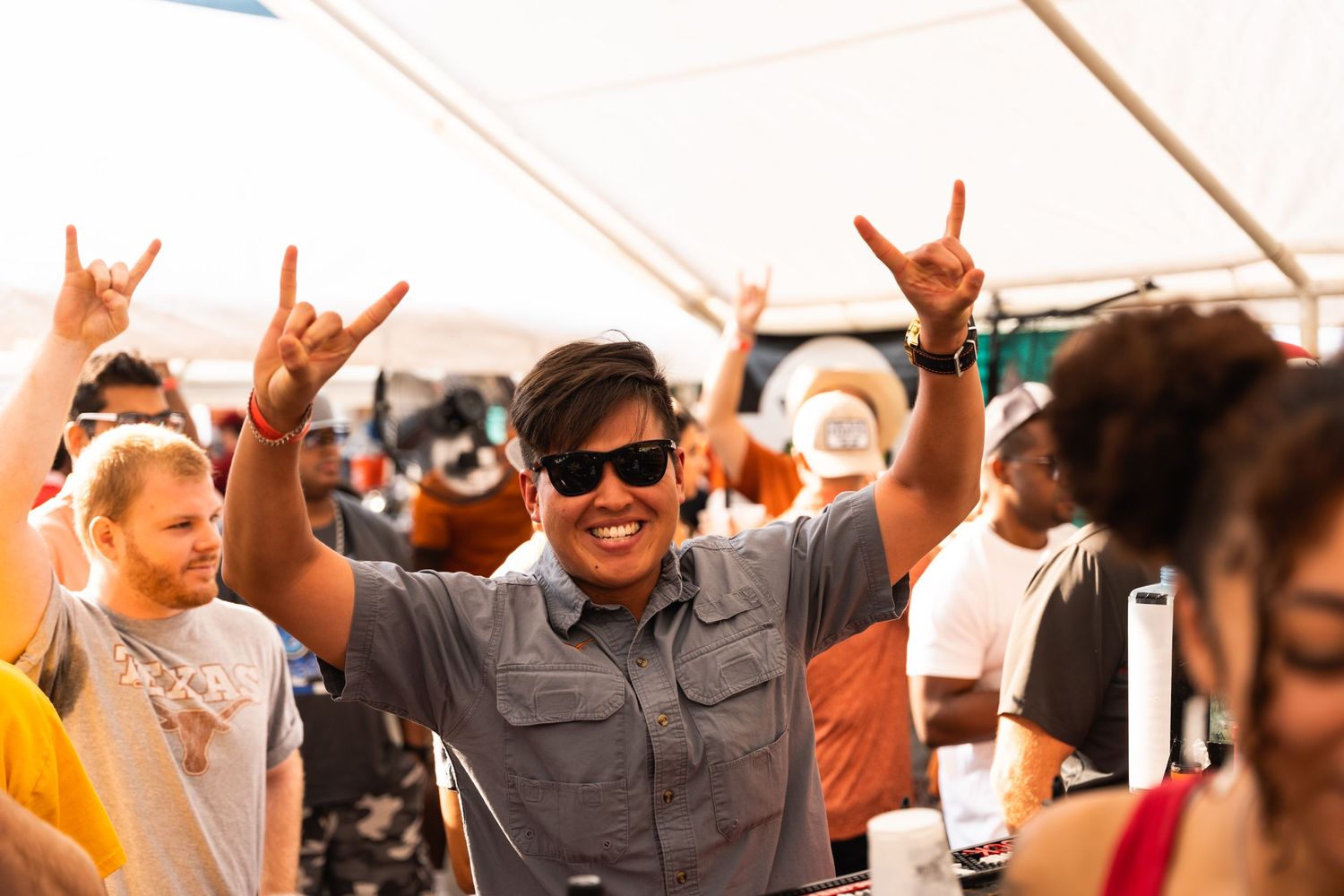 Excited partygoer in grey shirt and sunglasses throws up rock hand signs under festival tent.