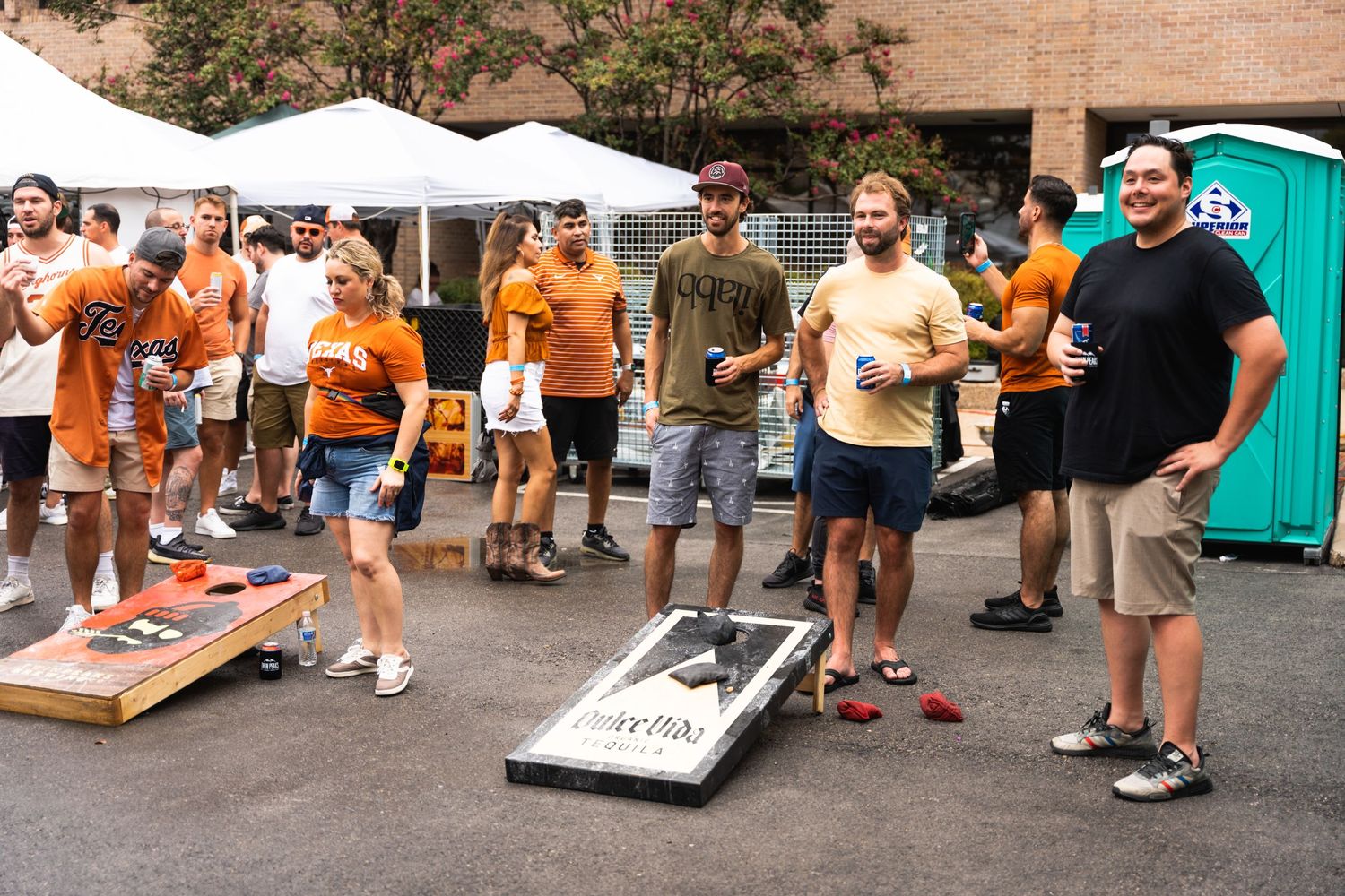 People playing cornhole games at an outdoor party with orange tents and portable restrooms.