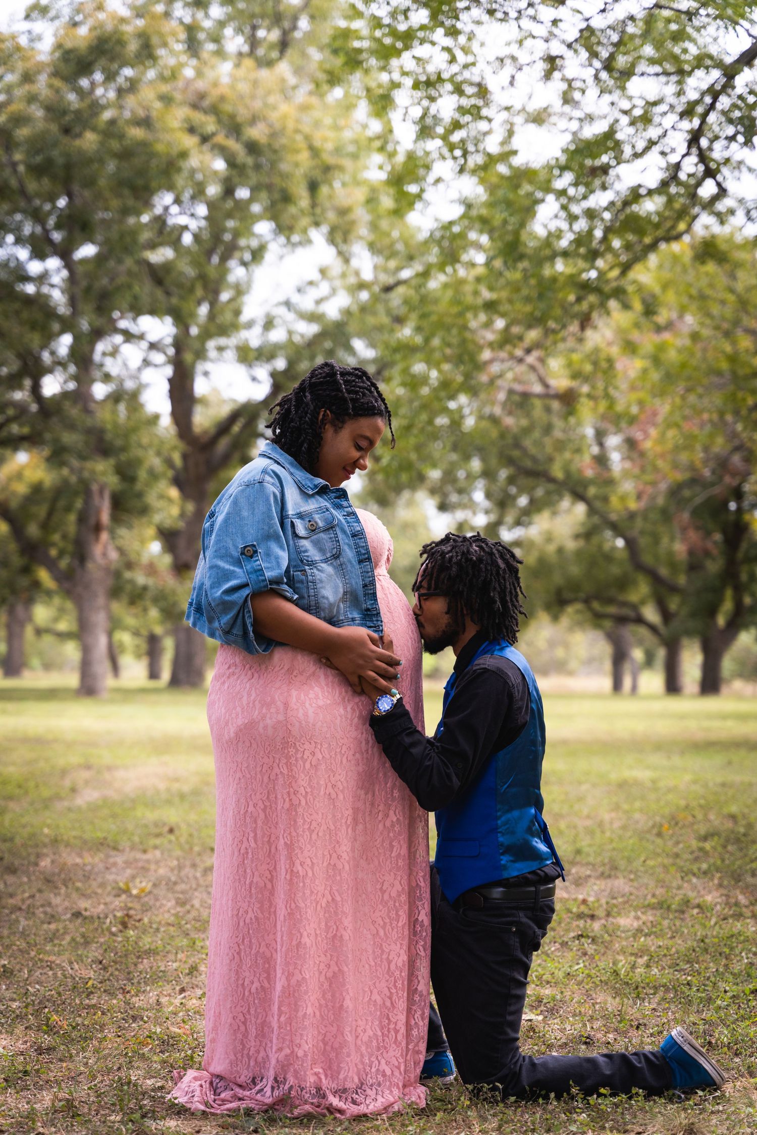 A tender pregnancy photo with the partner kneeling to embrace a growing baby bump in a pink dress outdoors.