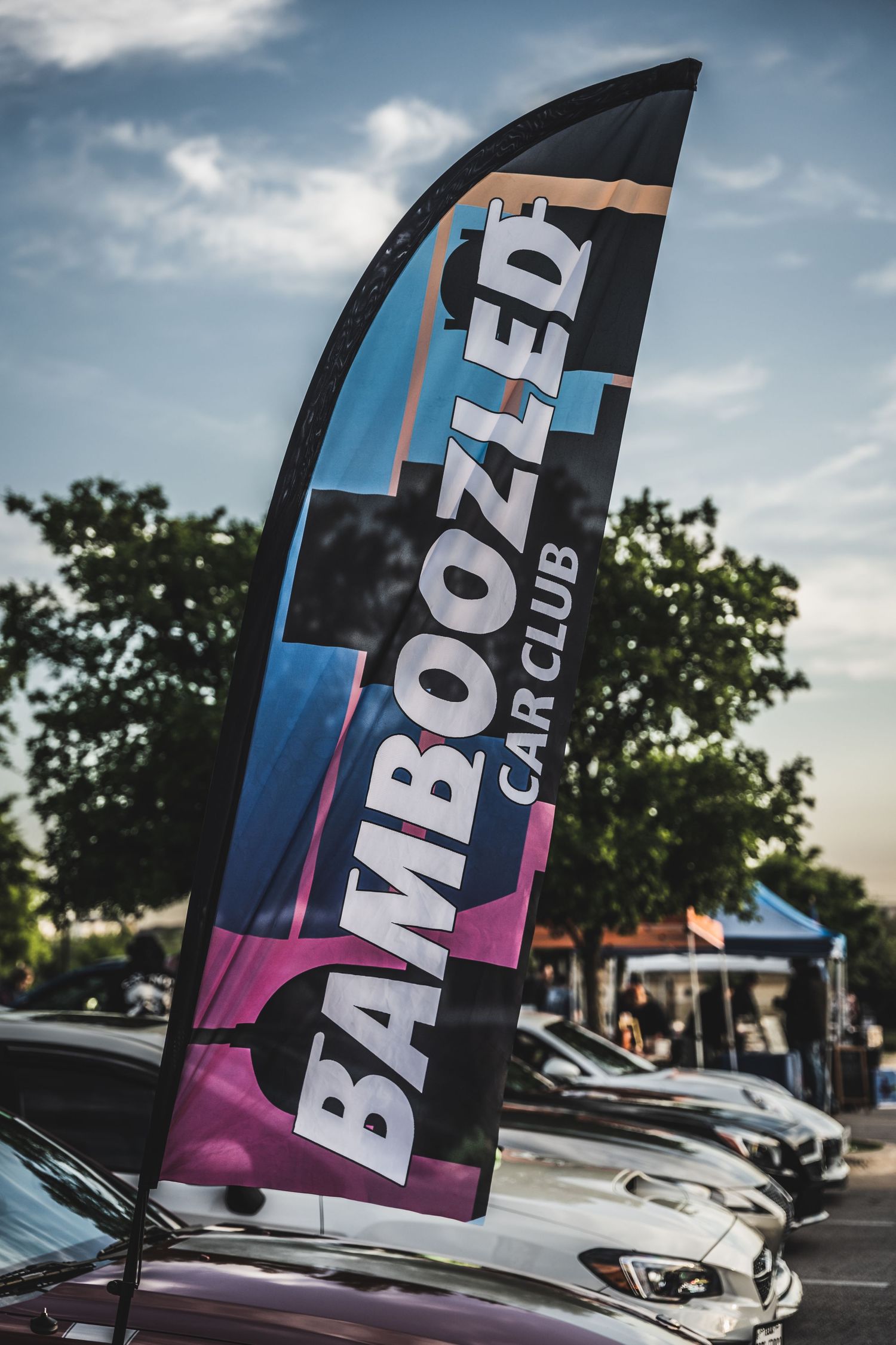 A tall Bamboozled Car Club banner flag against a blue sky at an auto show.
