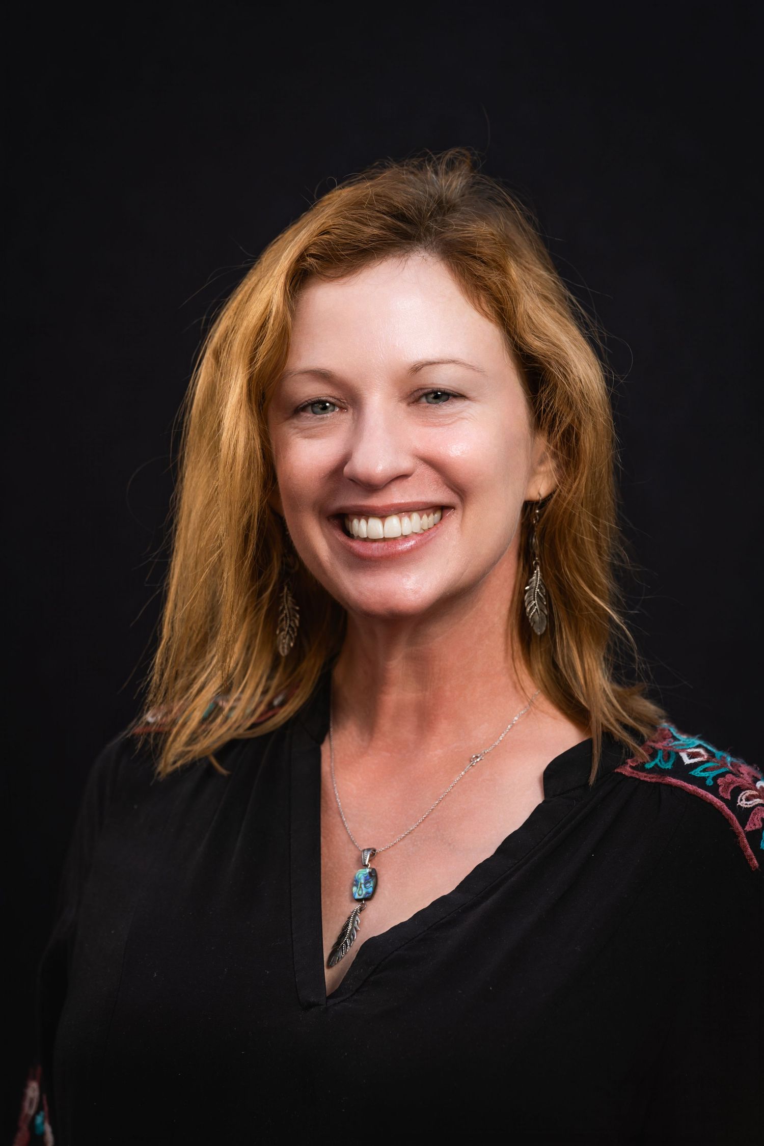 Professional headshot featuring person with auburn hair wearing black top and turquoise pendant necklace.