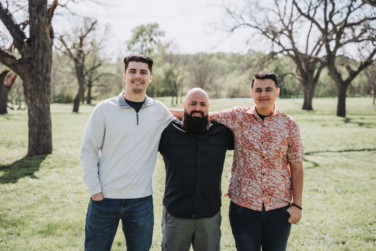 Three people standing together outdoors in a grassy field with trees behind them.