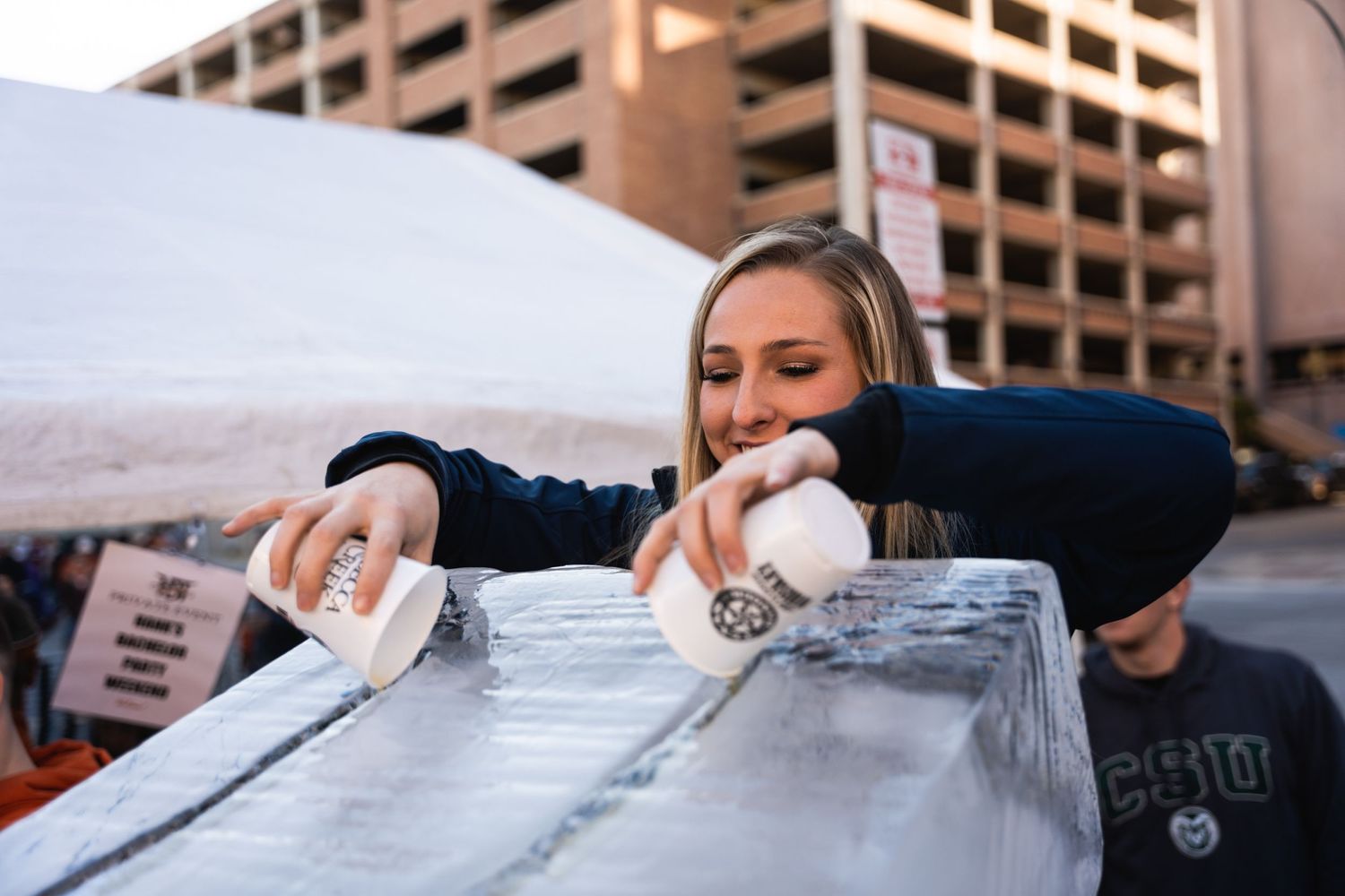 Someone pours liquid onto a large ice block at an outdoor event.