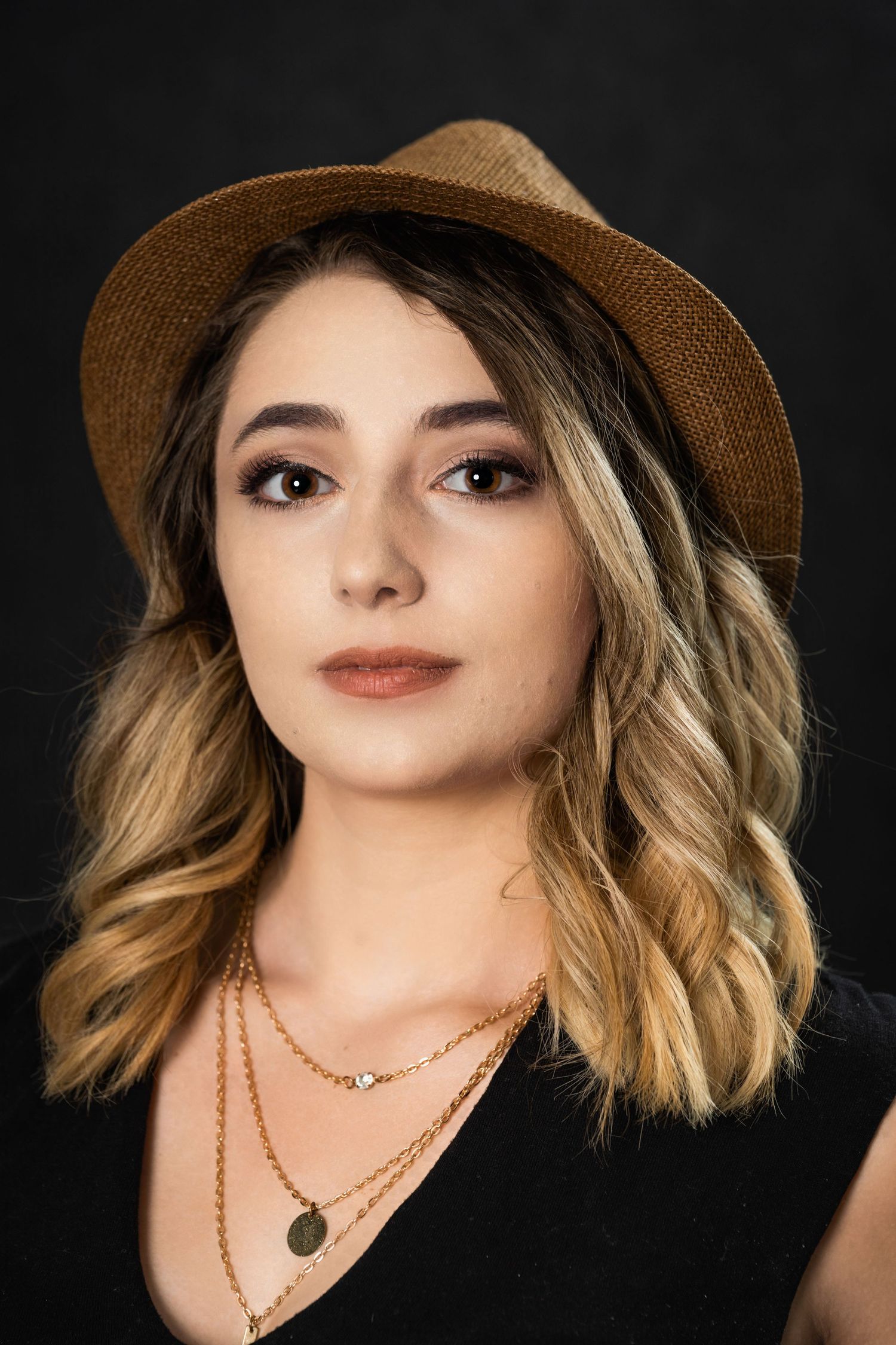 Stylish portrait with tan fedora hat and layered jewelry against dark backdrop.