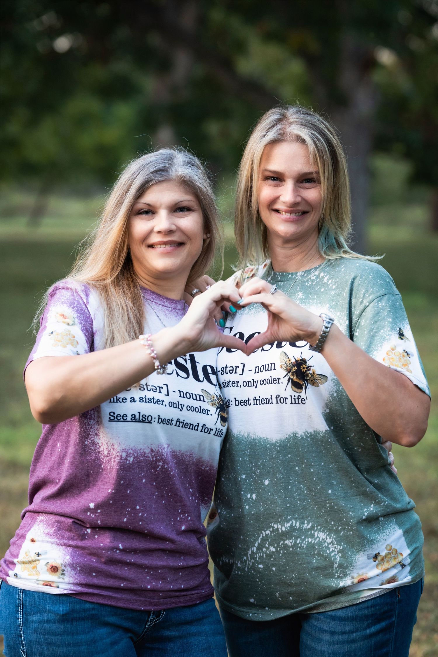 Two people in matching shirts make heart shapes with their hands while smiling outdoors.