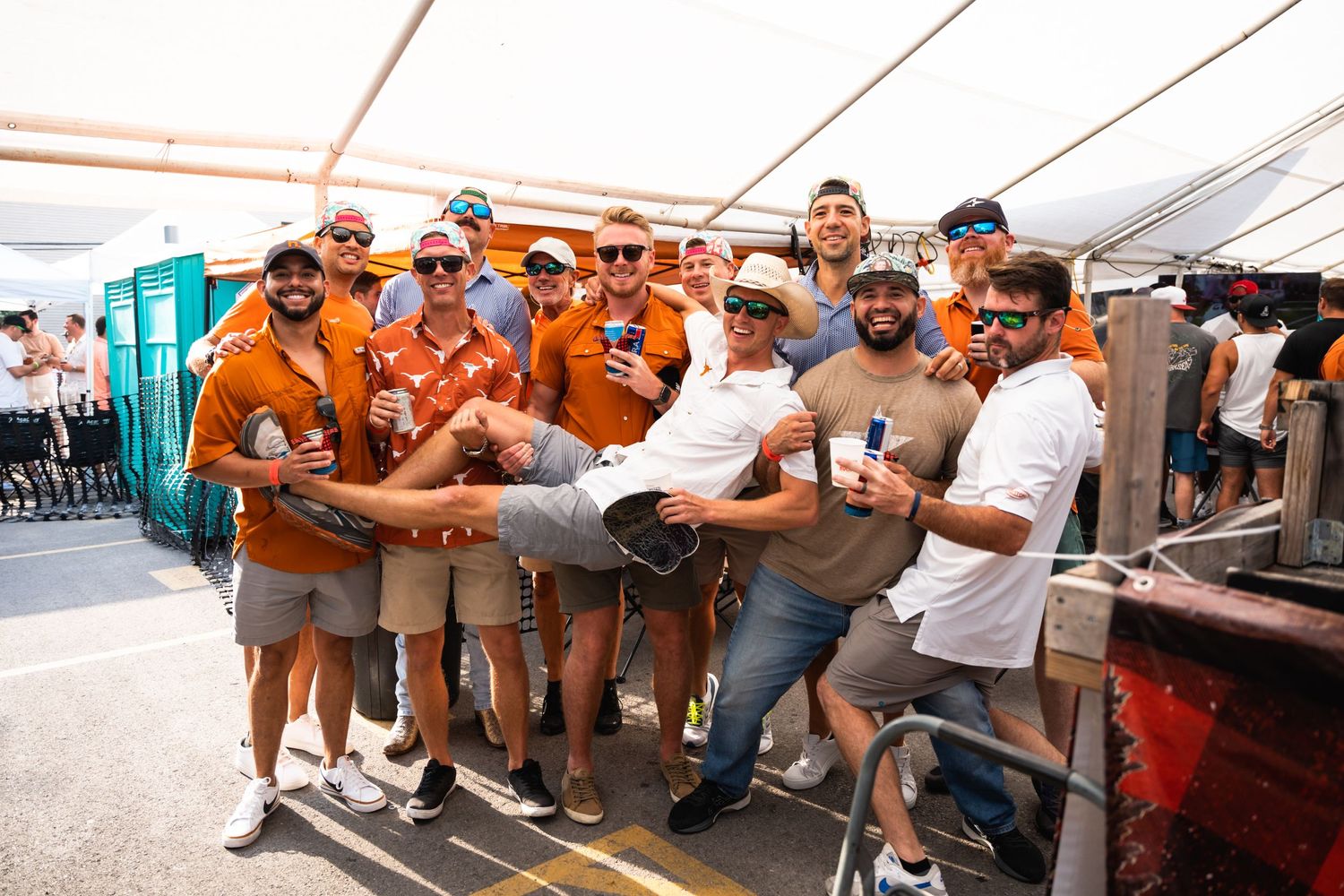 Group of people in orange attire celebrate together at outdoor event under white tent.