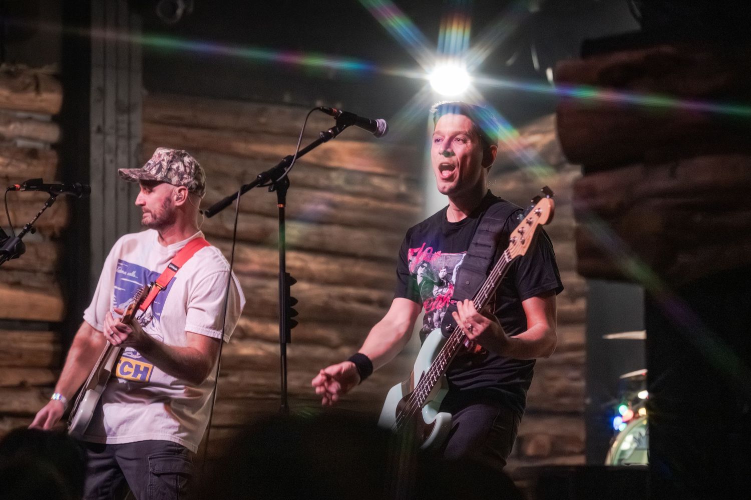 Musicians perform on stage with guitars and microphones in a dimly lit venue with wooden wall panels.