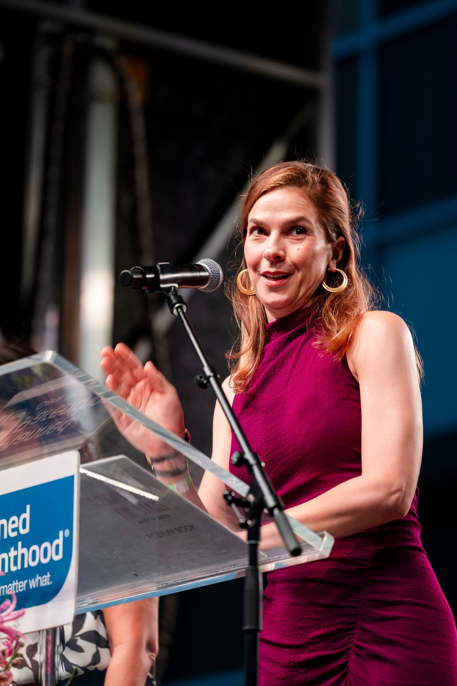 A speaker in a burgundy dress gestures animatedly while presenting at a podium with a microphone during an event.