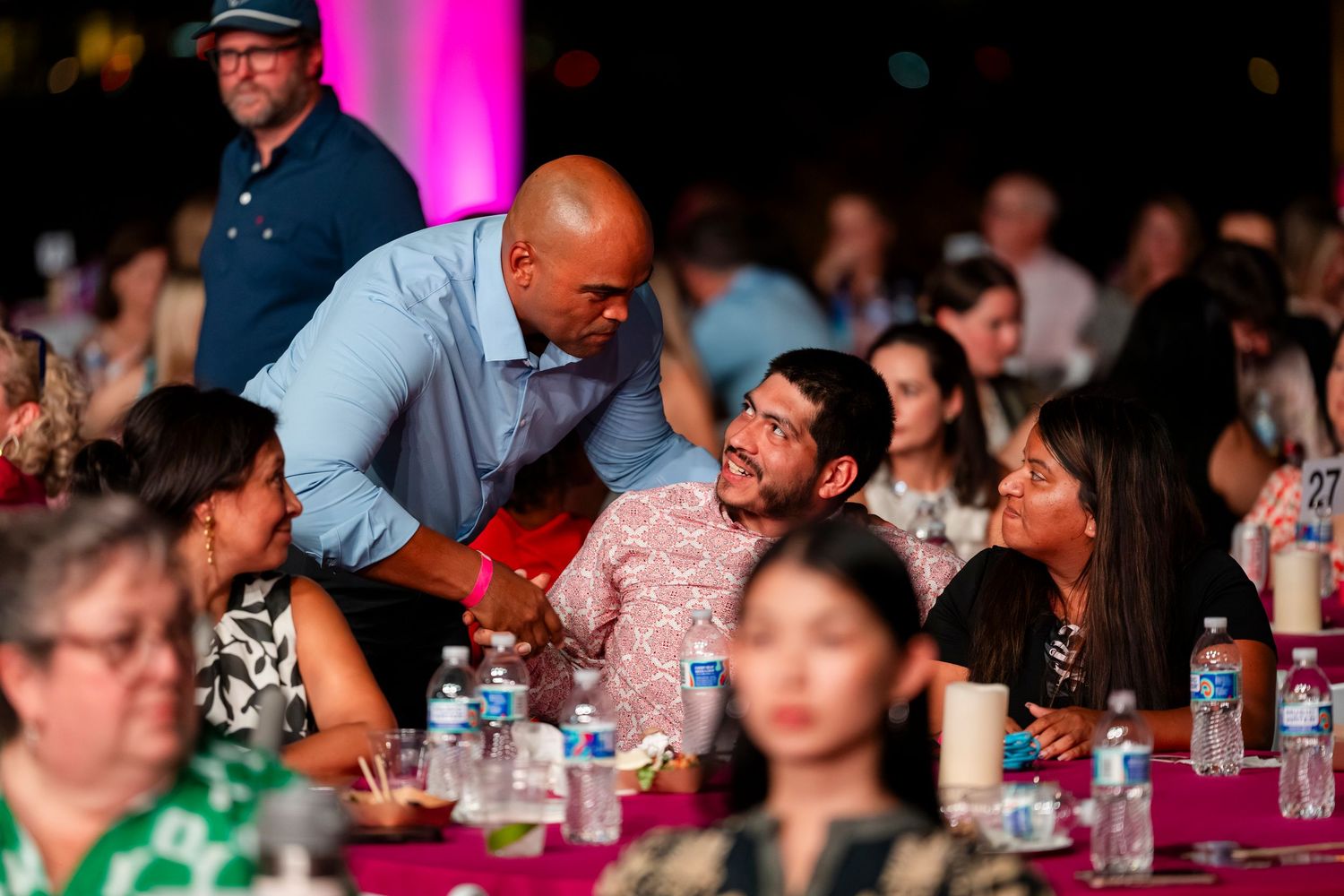 A group of people sitting at tables during an indoor event with pink and purple lighting in the background.
