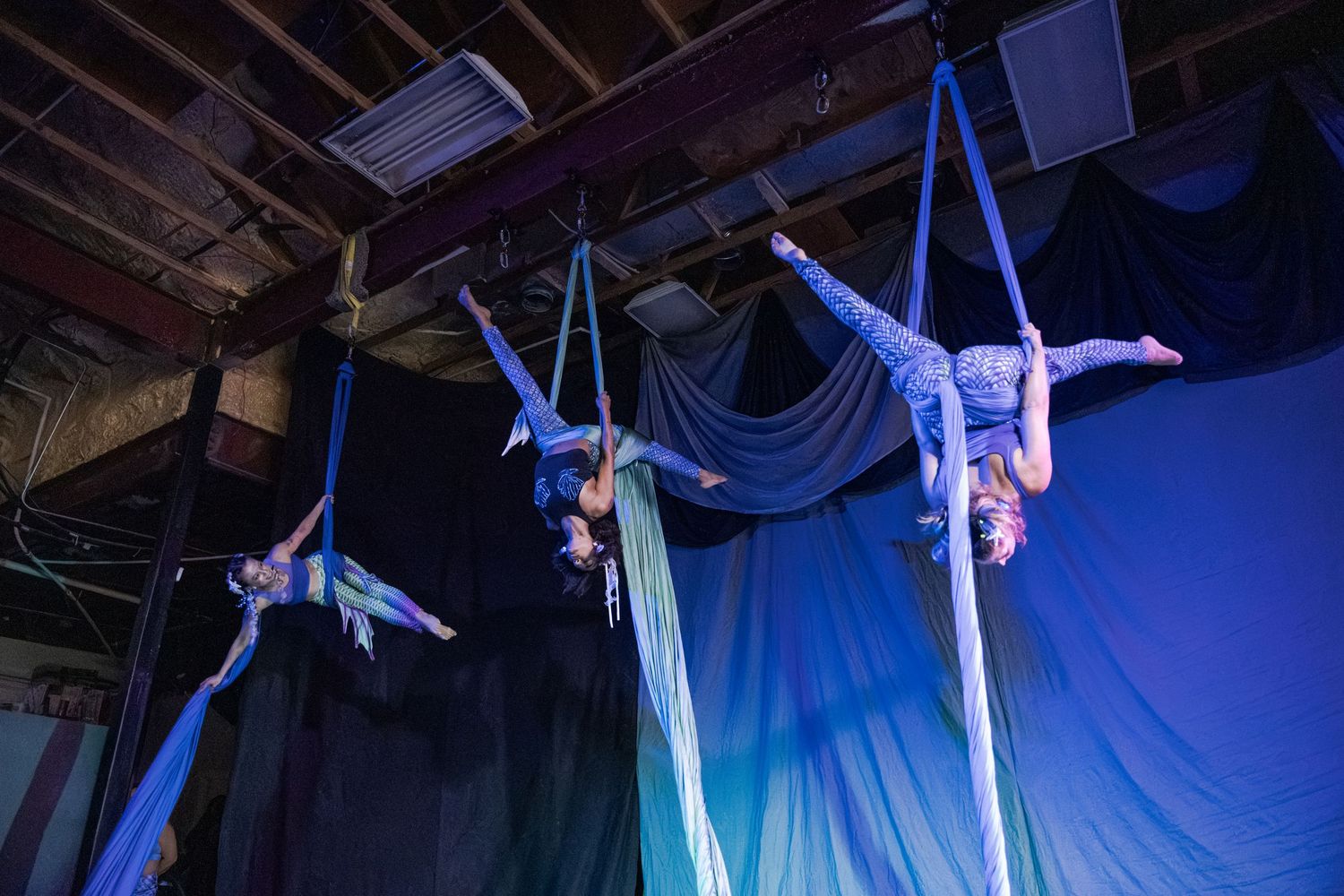 Three aerial silk performers suspended mid-air on blue fabric in a dramatic indoor circus performance under stage lighting.