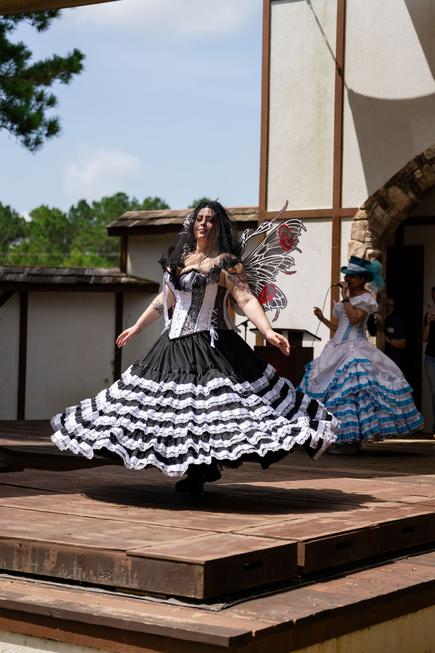 Woman in black and white ruffled fairy costume twirling on outdoor stage at renaissance festival.
