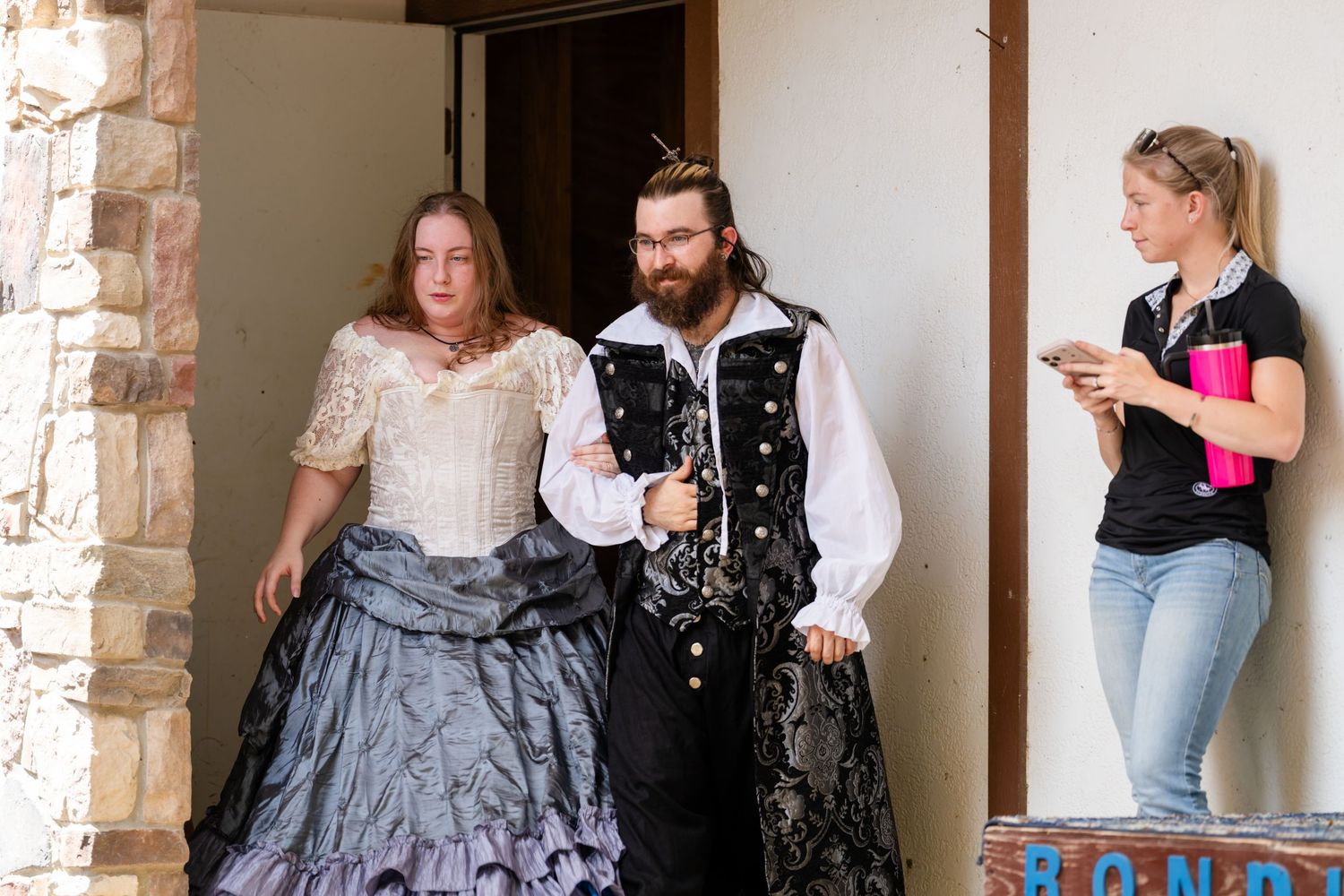 Two people in Victorian costumes exit a doorway while a woman in jeans checks her phone nearby.