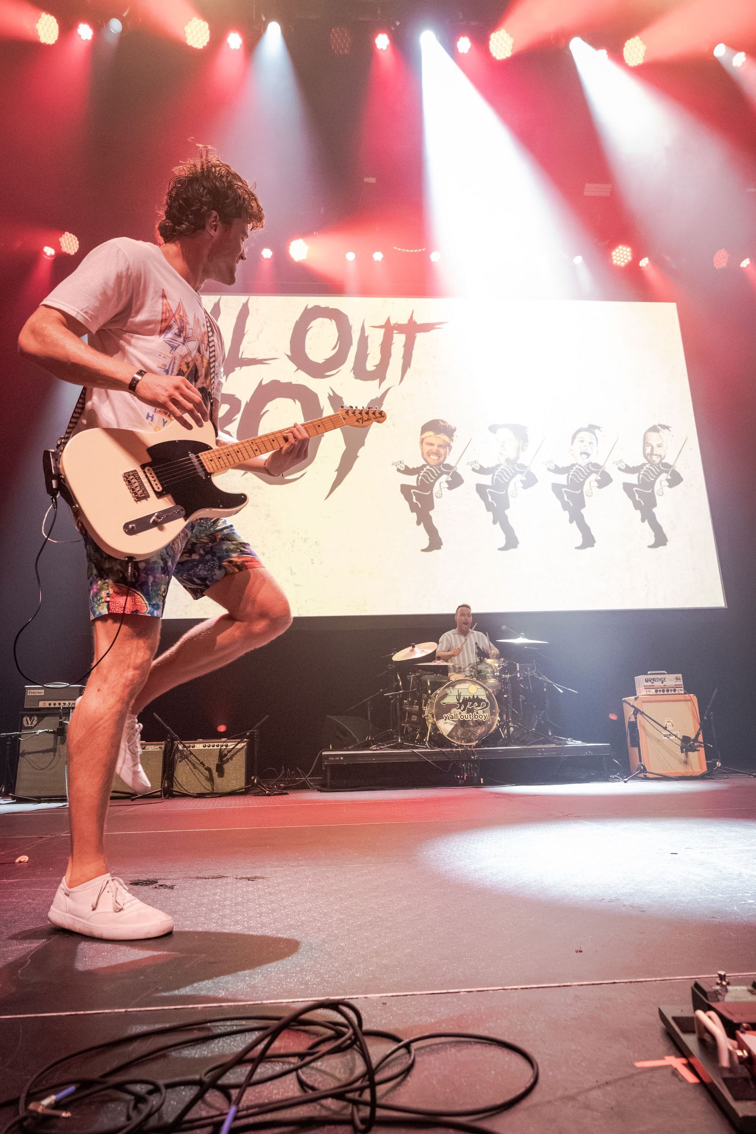 Guitarist performs energetically on stage at a Fall Out Boy concert with band logo backdrop and drummer visible behind.
