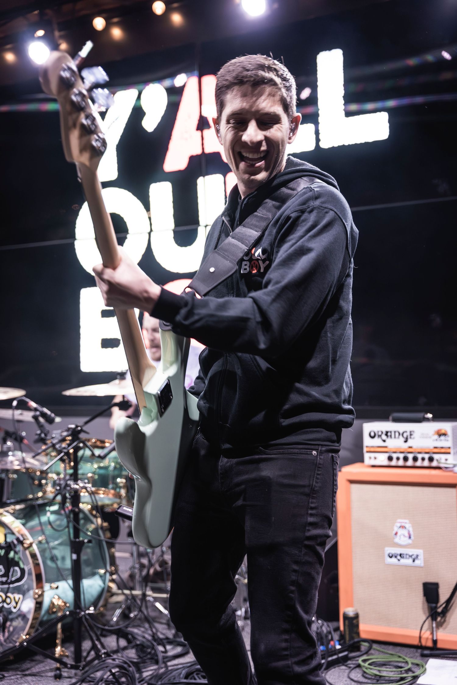 Guitarist performing on stage with 'Y'ALL OUT' signage and orange amp in background.