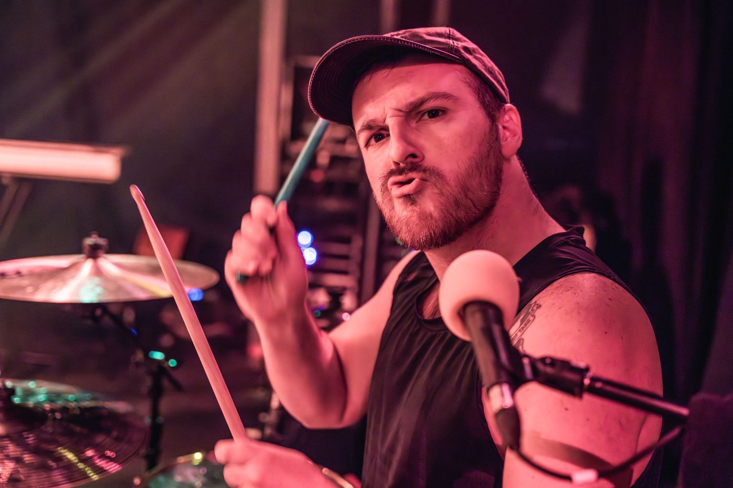 Drummer in black cap performing intensely under red stage lighting.
