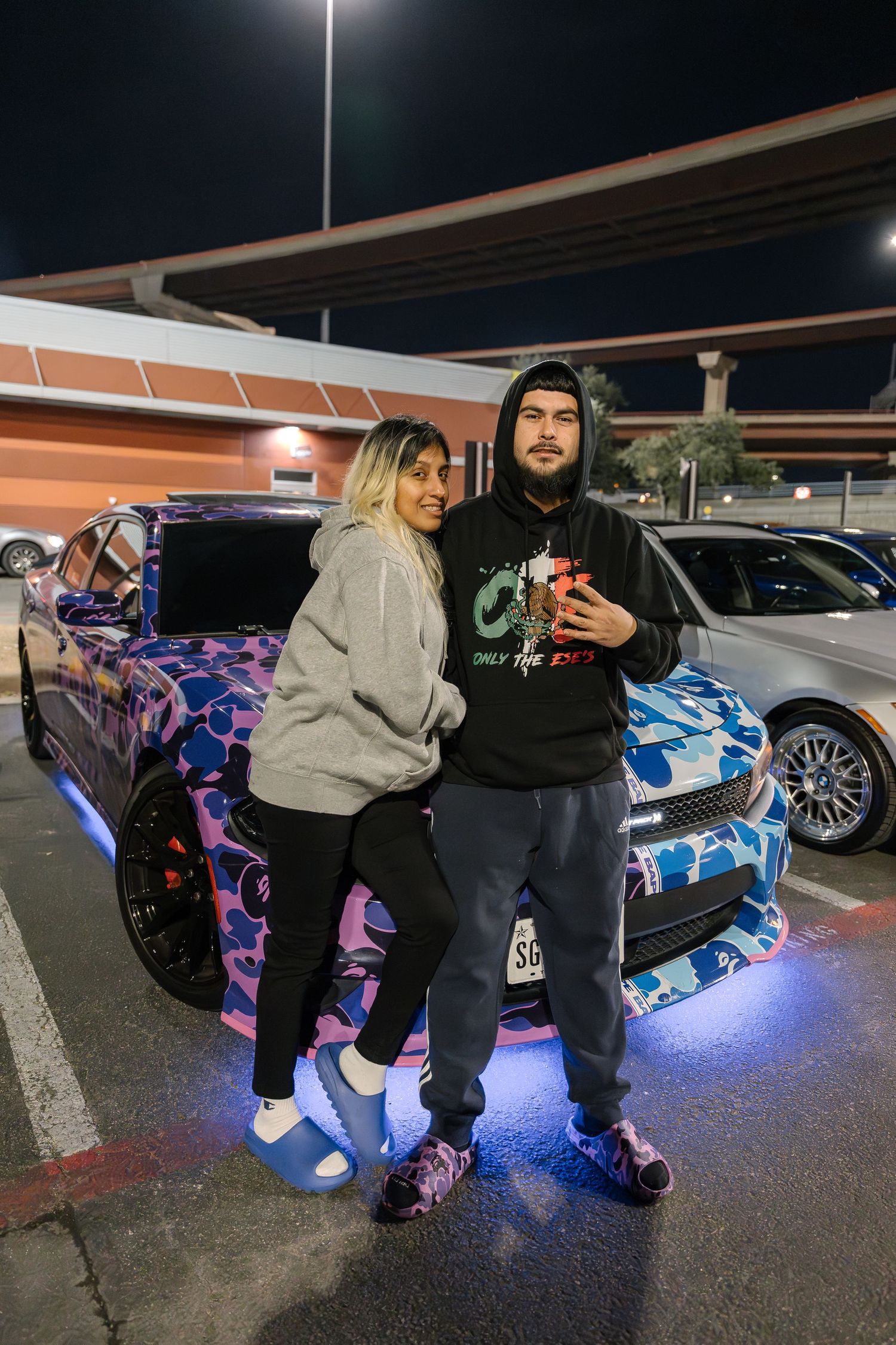 A couple stands in front of a car with custom blue LED lighting and camouflage wrap at night.