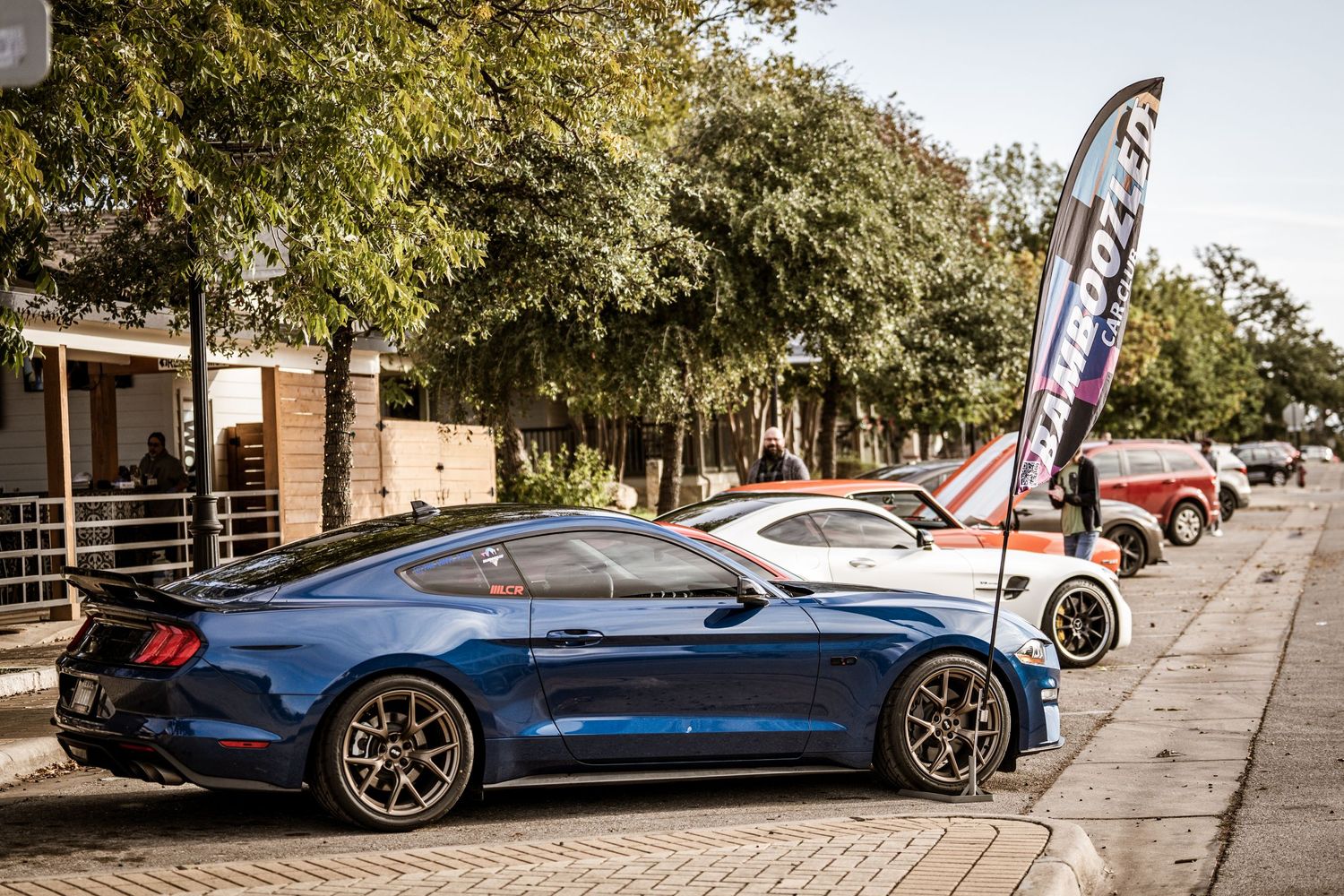 Line of luxury sports cars parked along a tree-lined street with event banners.