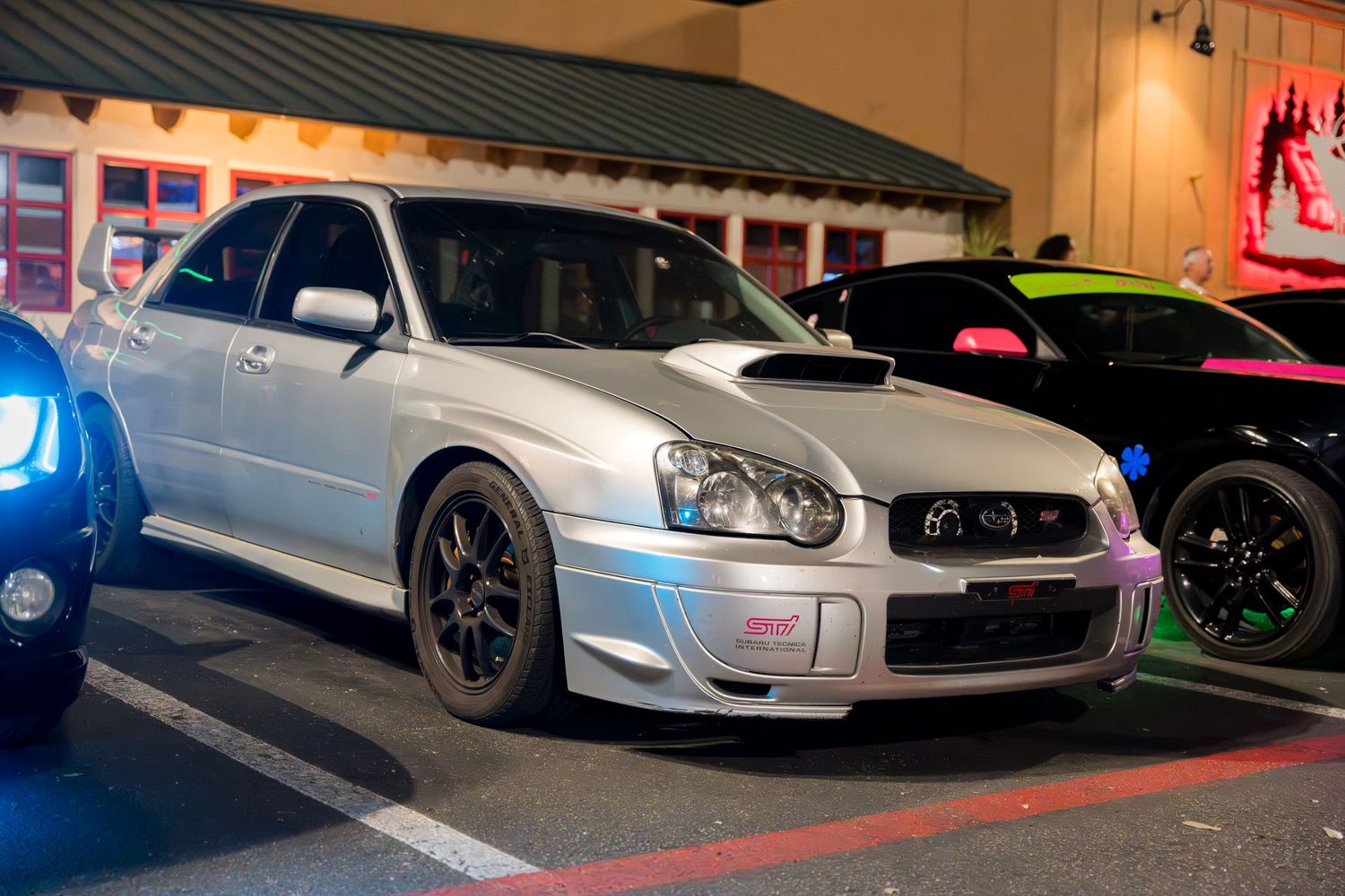 Modified silver Subaru WRX STI with custom wheels and blue lighting parked at night.