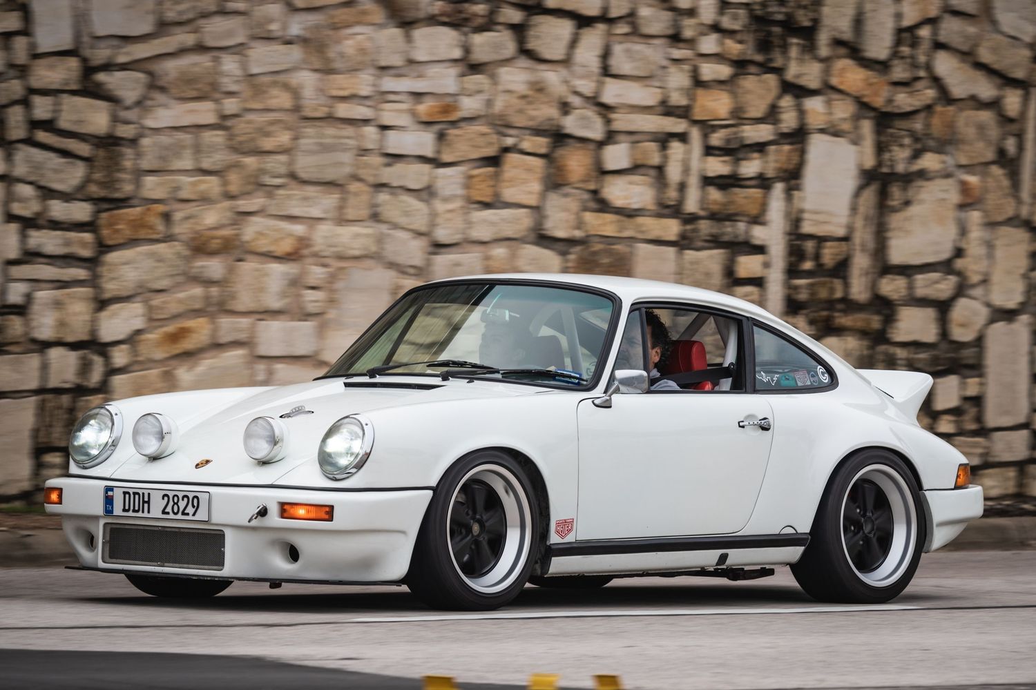 Classic white Porsche 911 parked in front of a stone wall featuring aftermarket wheels.