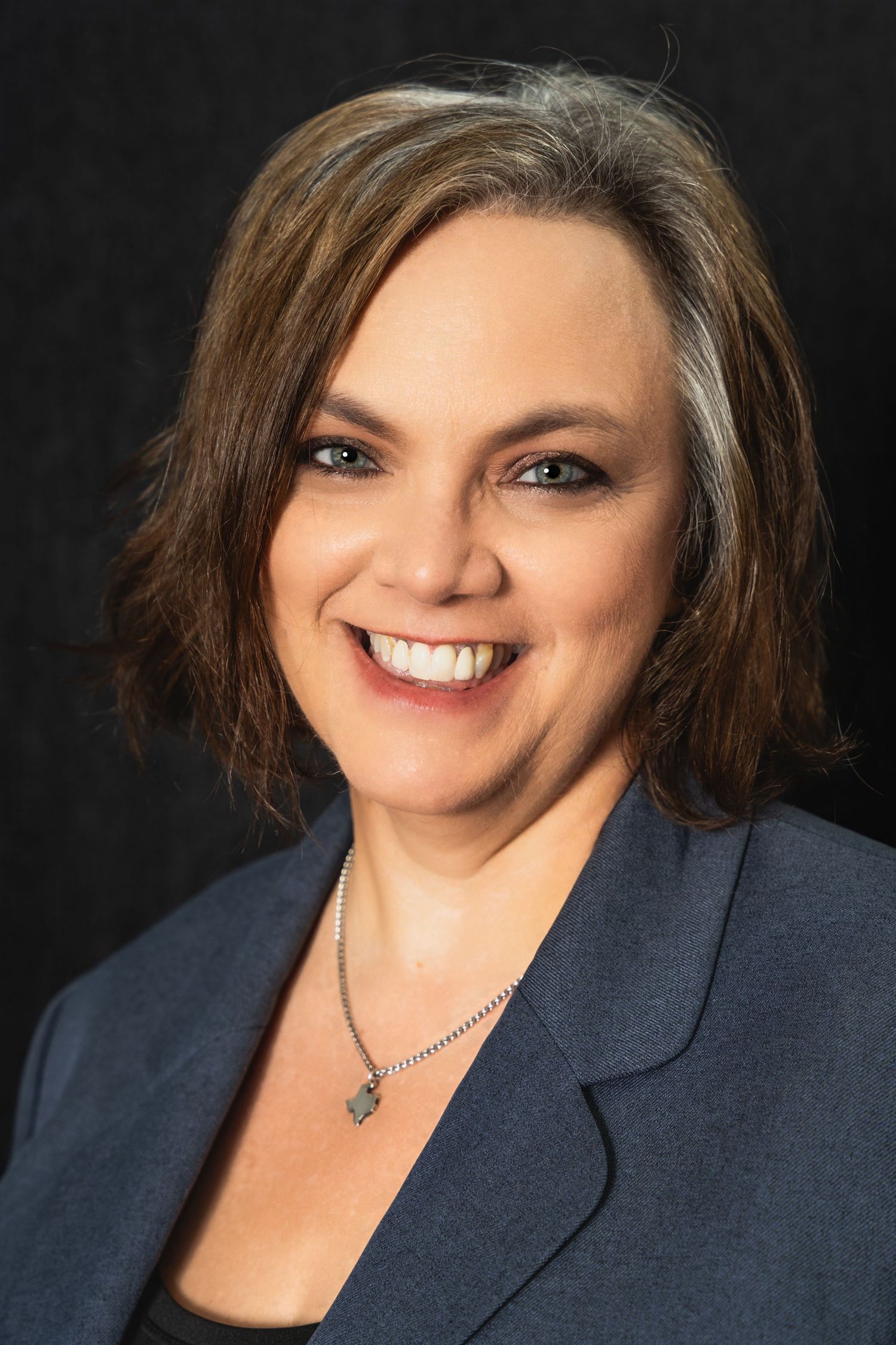 Professional headshot of a business professional wearing a navy blazer and silver necklace against dark background.