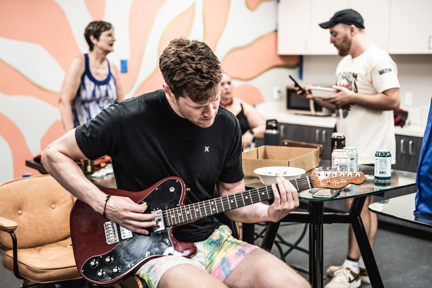 A musician plays an electric guitar in a studio room with a coral-colored abstract wall pattern.