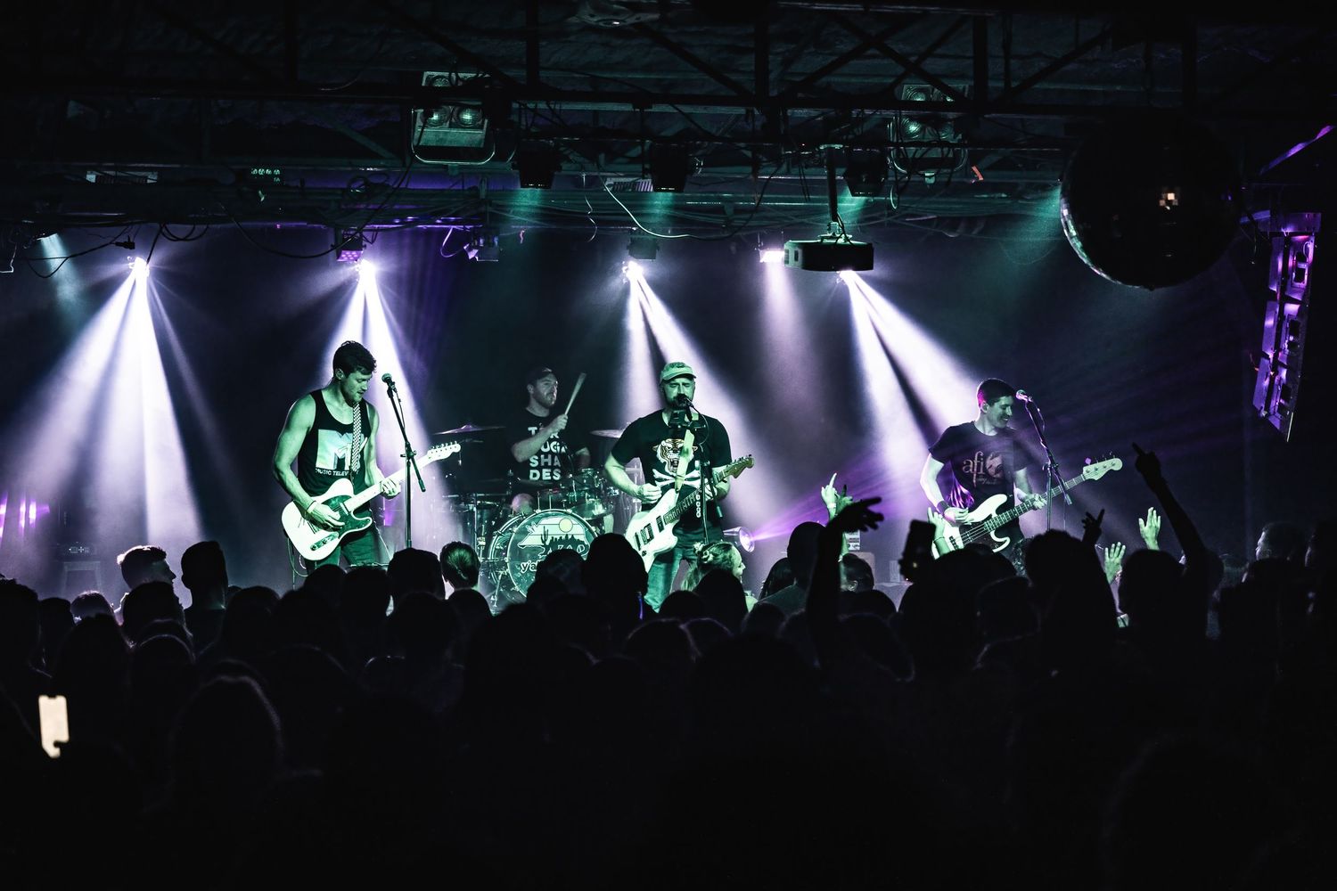 Concert venue filled with silhouetted crowd watching performers under dramatic white spotlights.