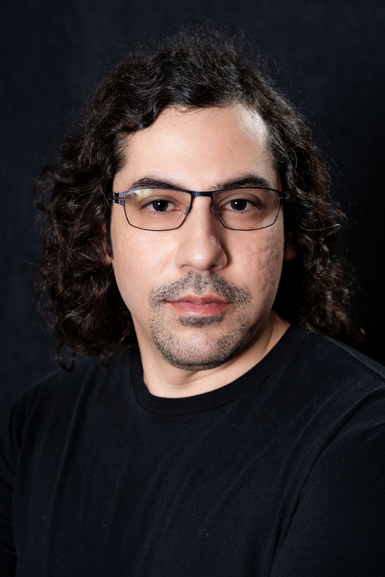 Professional studio portrait of a person with curly hair wearing glasses and black shirt against dark background.