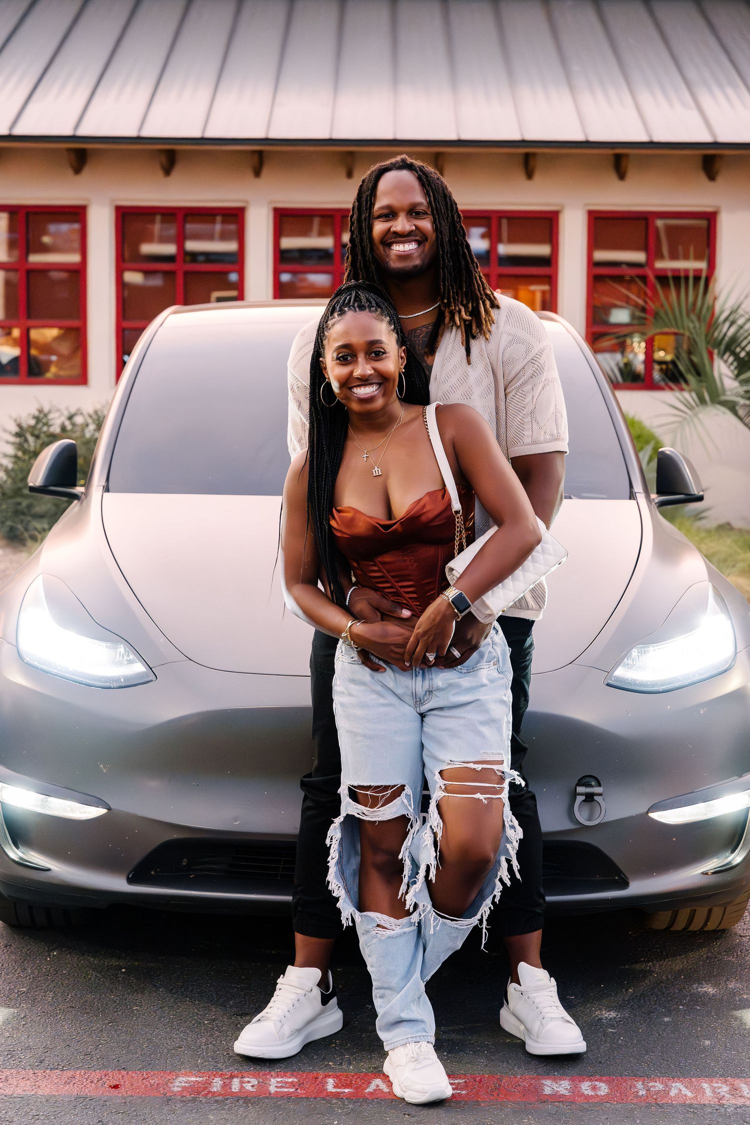 Happy couple posing in front of a silver Tesla Model 3 electric vehicle.