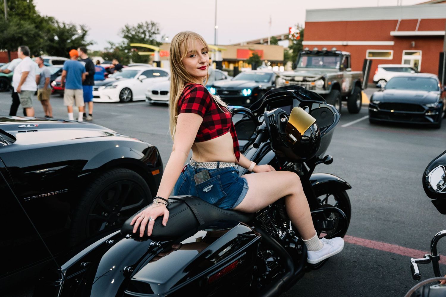 Person posing on black motorcycle at outdoor car meet.