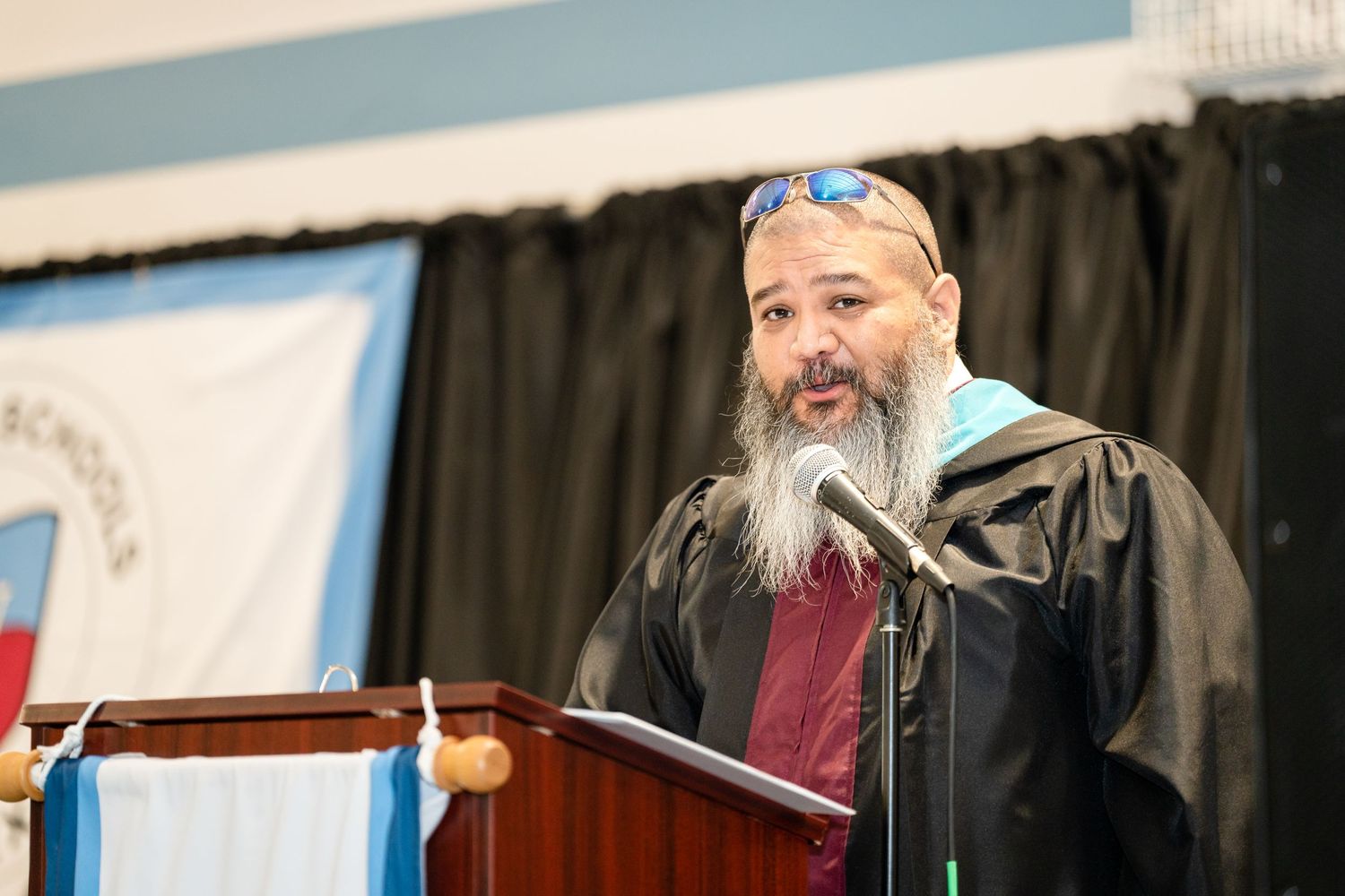 Speaker in academic regalia with gray beard addresses audience at graduation podium.
